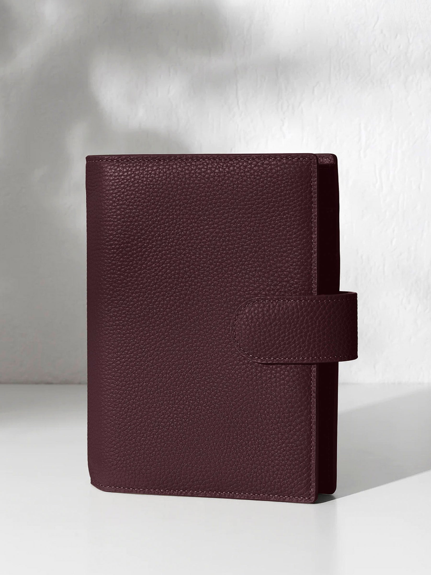 A closed dark burgundy leather planner with a closure displayed against a shadowed background.