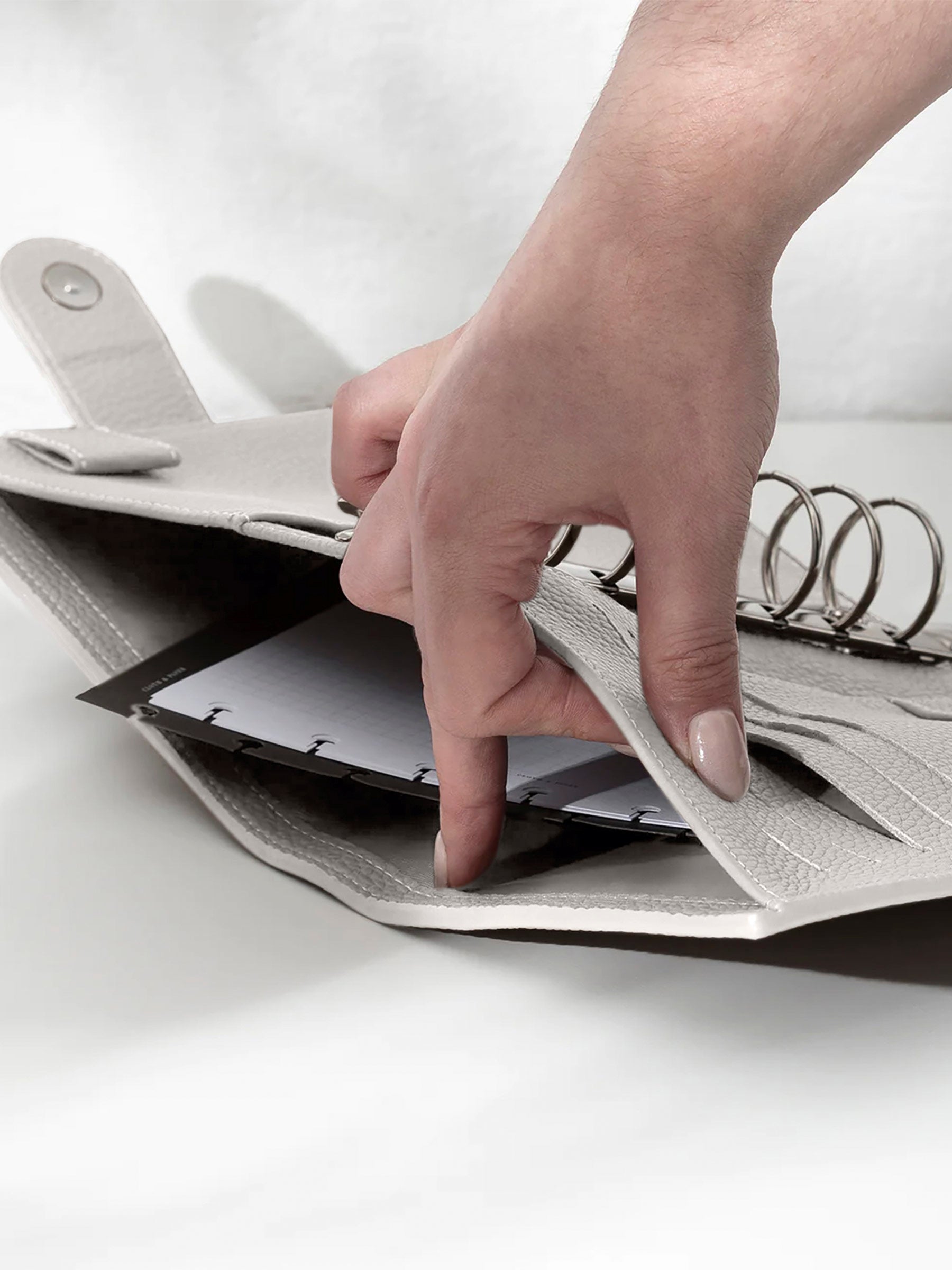 A fair-skinned hand shown opening the back pocket of the ash leather agenda on a off-white background. There are inserts and a dashboard inside the pocket.