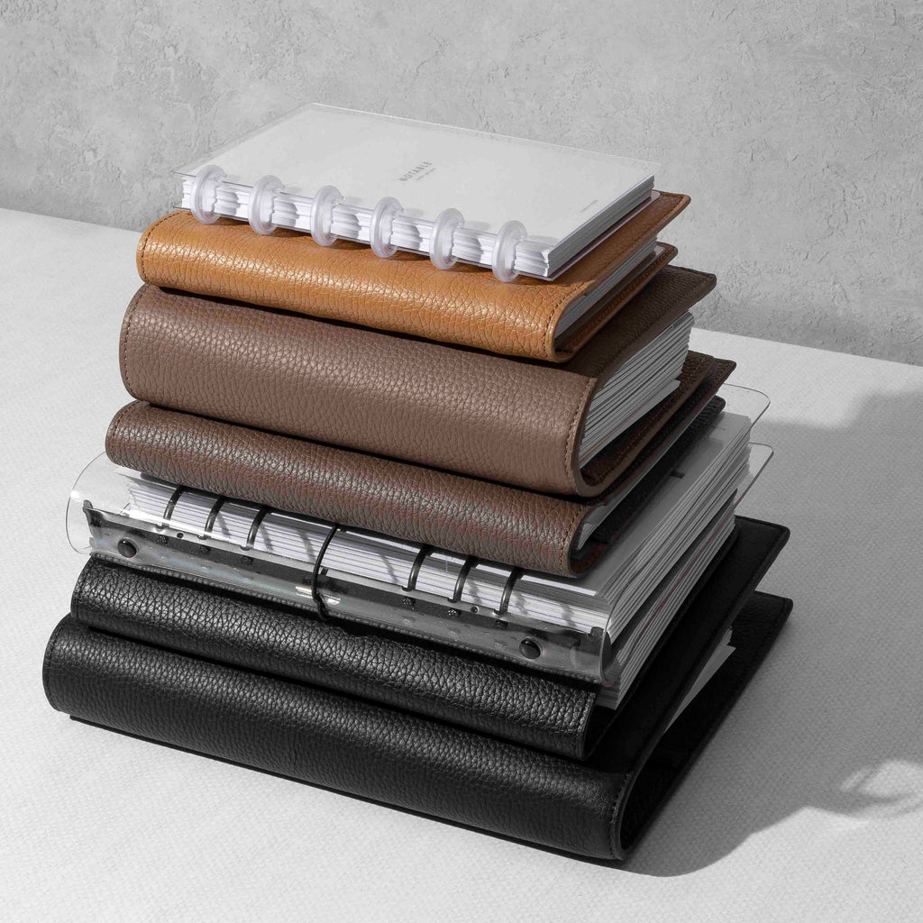 A stack of various Planners on a white desk including Leather Folios, Leather Agendas, Clear Vinyl, and Discbound.