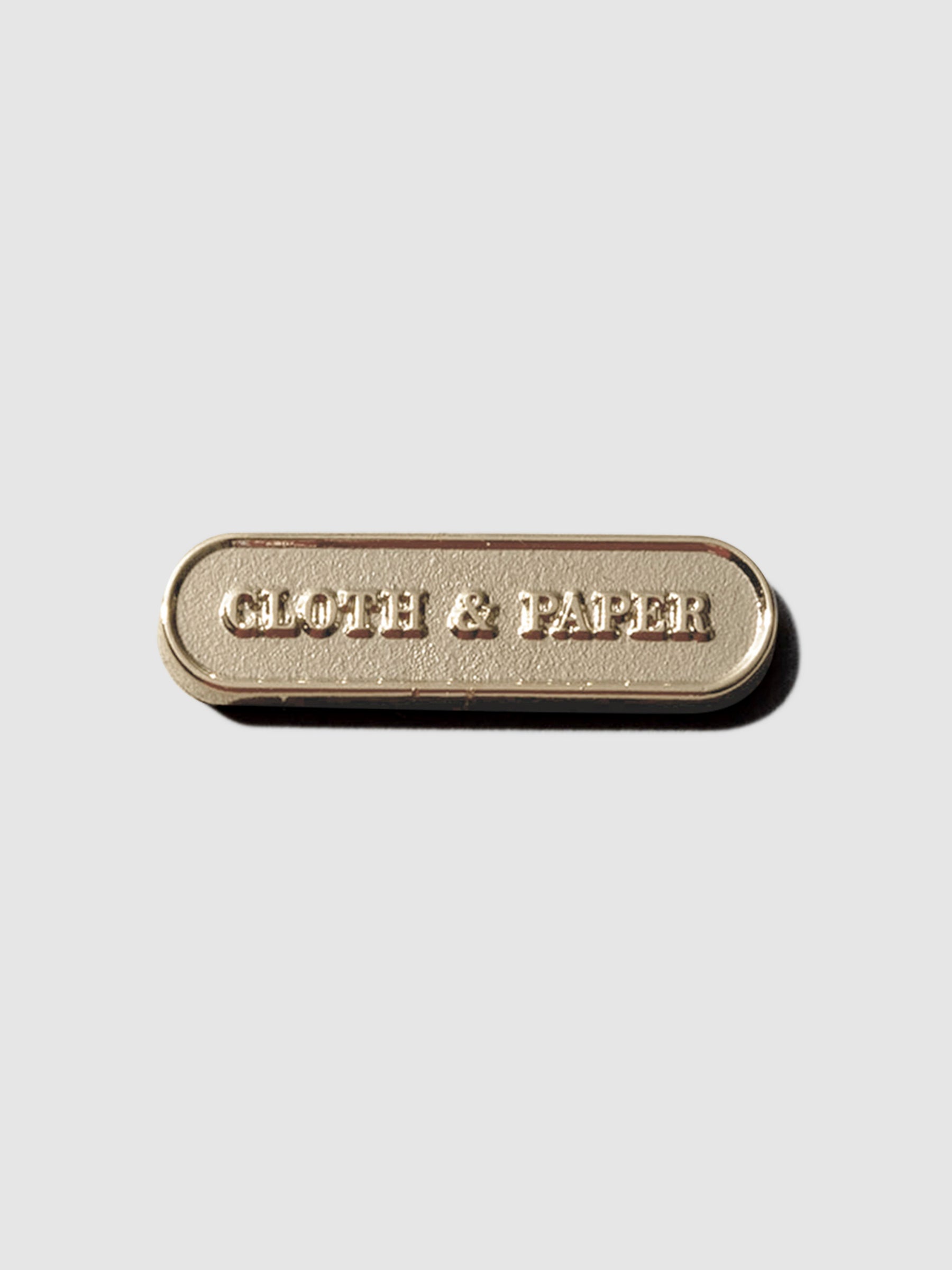 Gold pin displayed on a neutral background. Pin is pill shaped and has the Cloth and Paper logo inside of it.