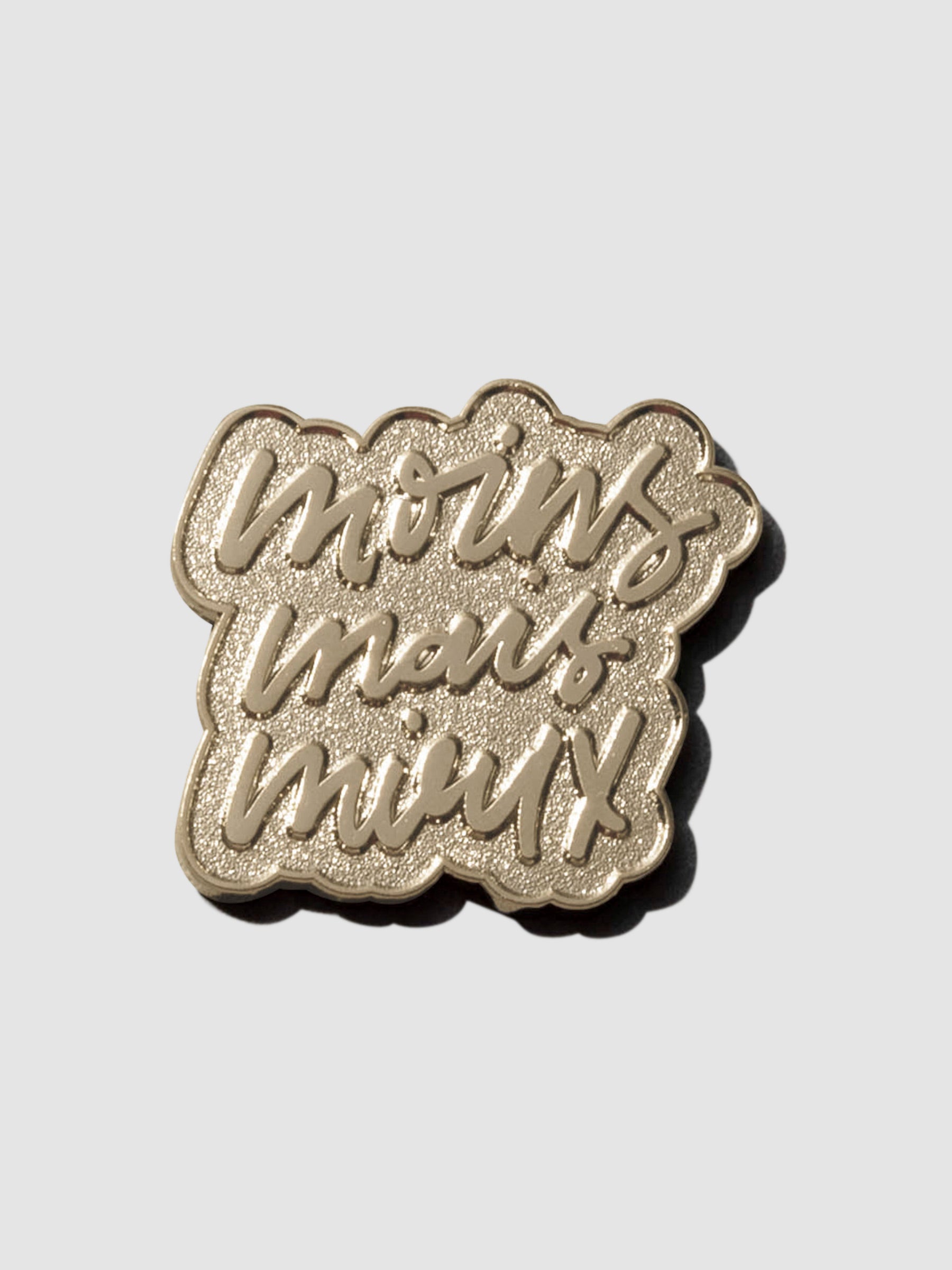 Gold pin displayed on a neutral background. Pin is handwritten text reading "moins mais mieux"