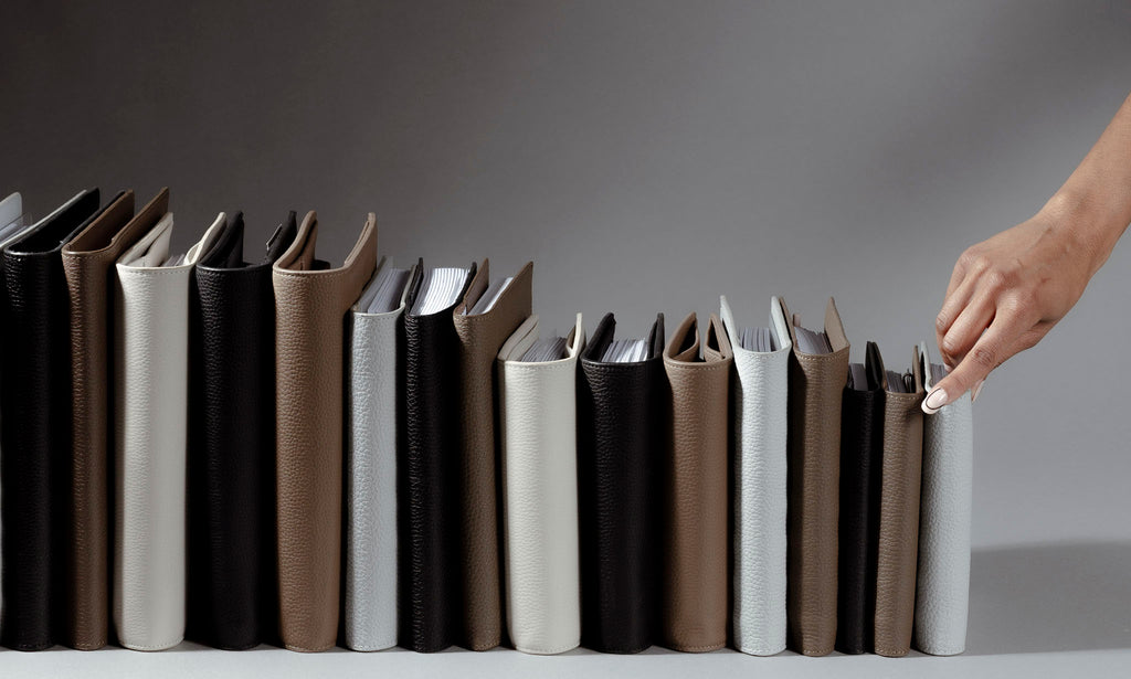 An image representing folios from our new Heirloom Leather Collection.  A hand is reaching for a Folio in color Veleta. The different folios and colors are standing up side by side with their spines facing out.