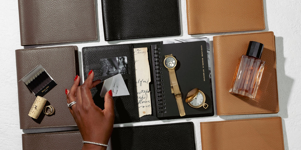 an image representing folios from our new Heirloom Leather Collection.  A hand is going through the contents of a planner with everyday accessories surrounding.
