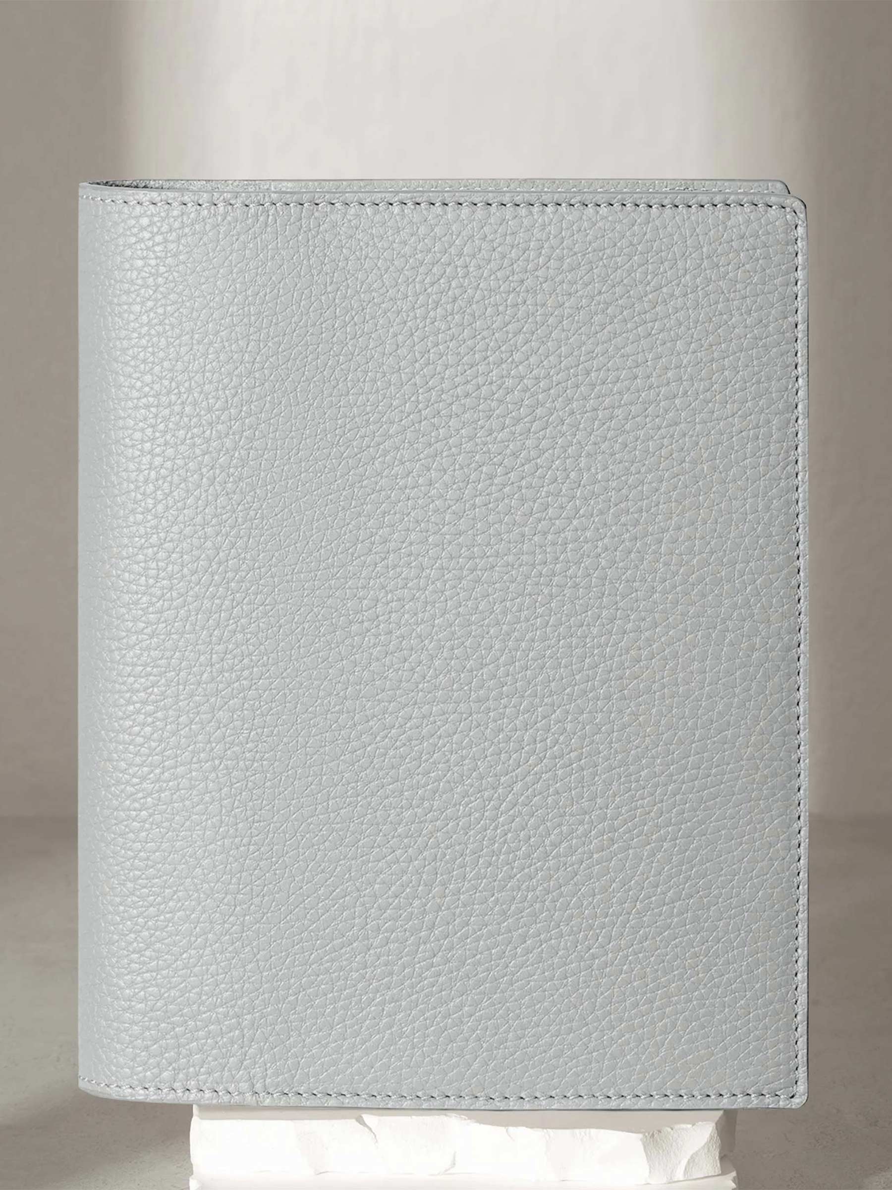 Heirloom Leather Folio, HP Classic, Veleta, Cloth and Paper. Veleta folio displayed on a white stone pedestal. The background is a natural textured off-white material, and a spotlight behind the folio highlights its placement.