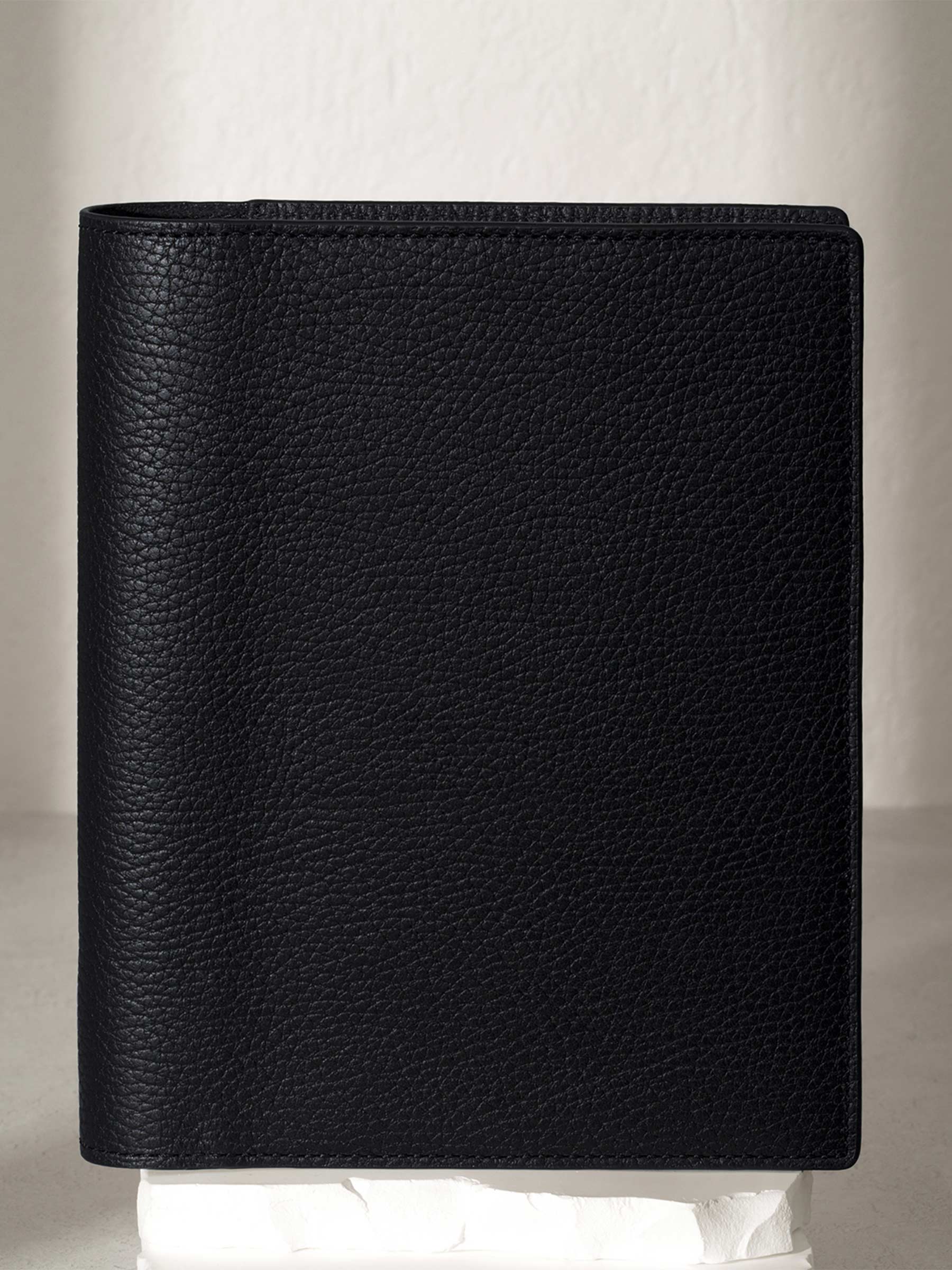 Heirloom Leather Folio, Small, Cloth and Paper. Padrillo folio displayed on a white stone pedestal. The background is a natural textured off-white material, and a spotlight behind the folio highlights its placement.