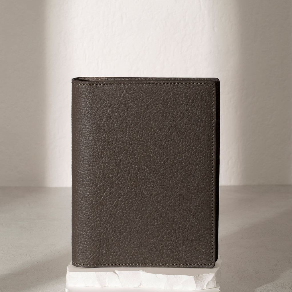 CP Petite Leon folio displayed on a white stone pedestal. The background is a natural textured off-white material, and a spotlight behind the folio highlights its placement.