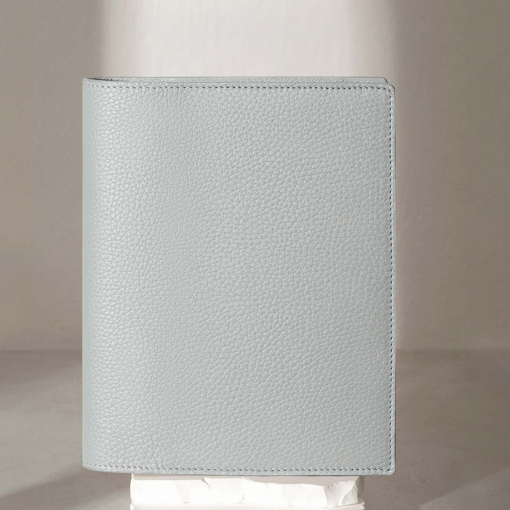 Heirloom Leather Folio, HP Classic, Veleta, Cloth and Paper. Veleta folio displayed on a white stone pedestal. The background is a natural textured off-white material, and a spotlight behind the folio highlights its placement.