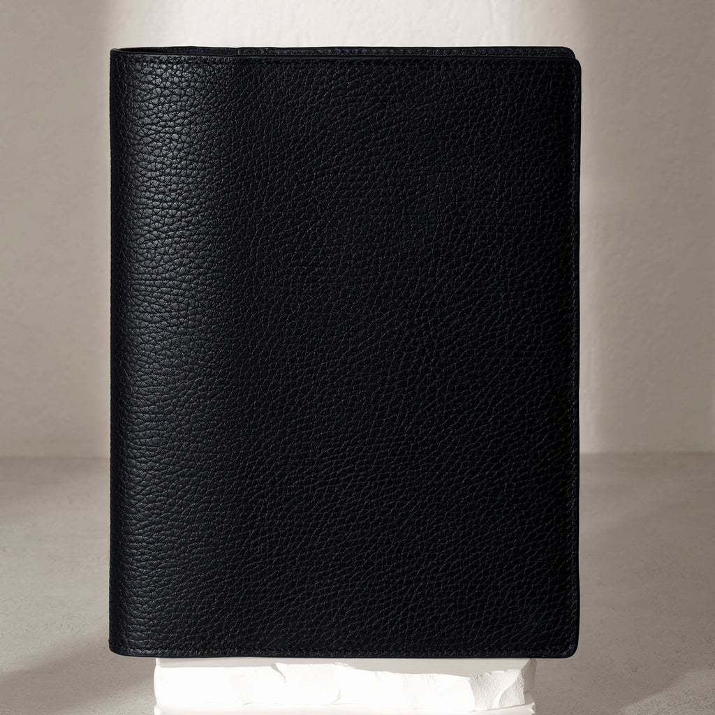 Heirloom Leather Folio, Large, Cloth and Paper. Padrillo folio displayed on a white stone pedestal. The background is a natural textured off-white material, and a spotlight behind the folio highlights its placement.