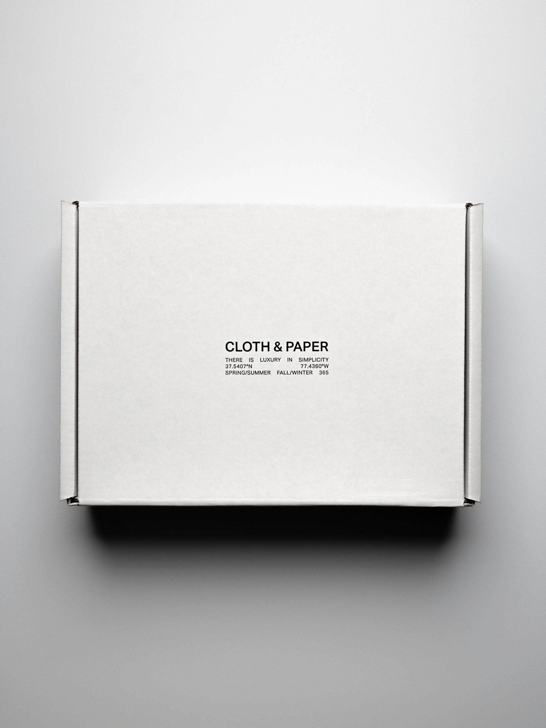 A image of the Intention Box. A white, sealed box, viewed from the top down on a neutral background. In black lettering on the top reads "Cloth & Paper. There is luxury in simplicity".