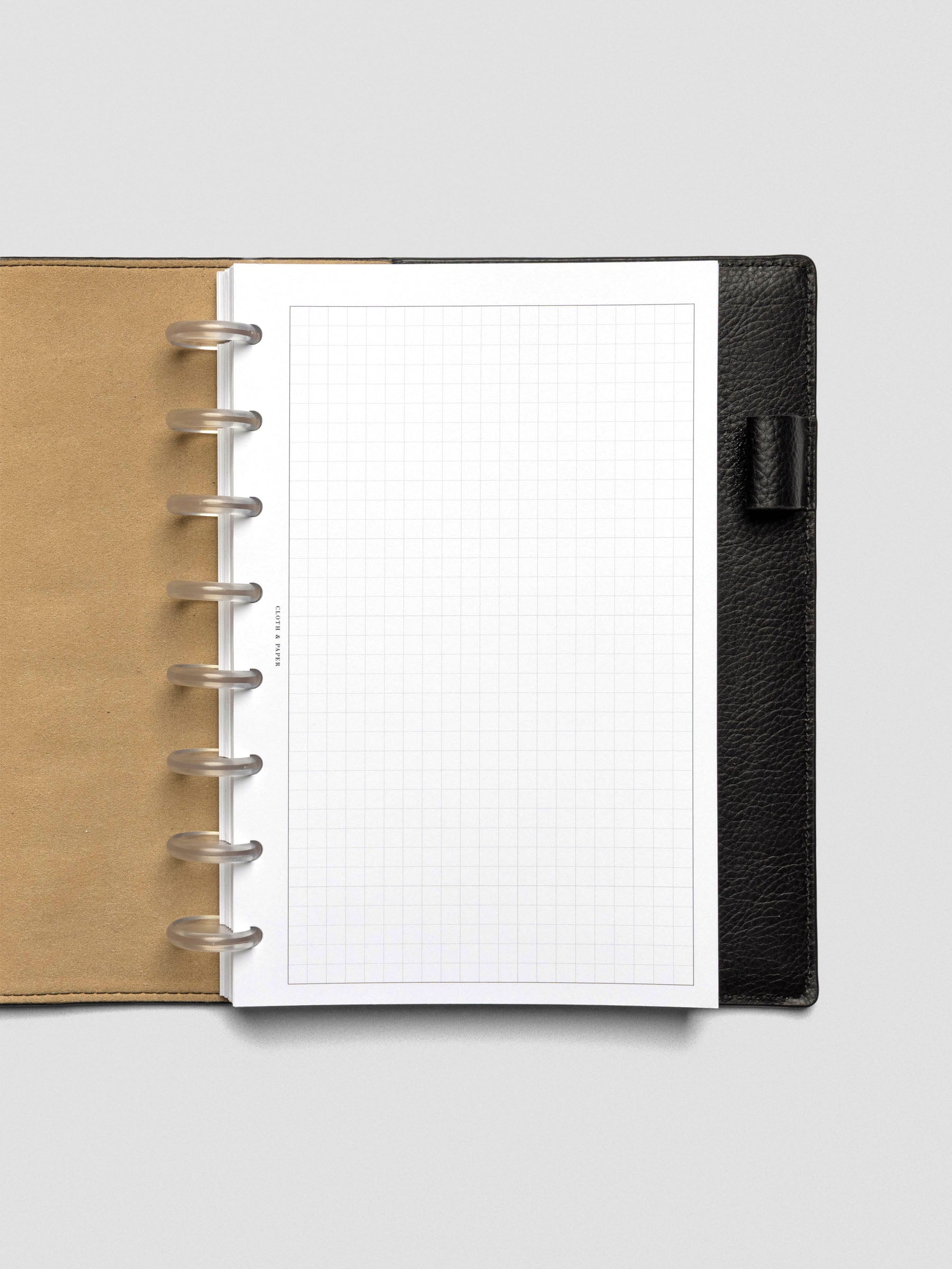 A single sheet of grid paper insert with a clear border, punched with 8-disc holes, placed inside a black leather planner with tan interior lining. The layout is minimal and unmarked.