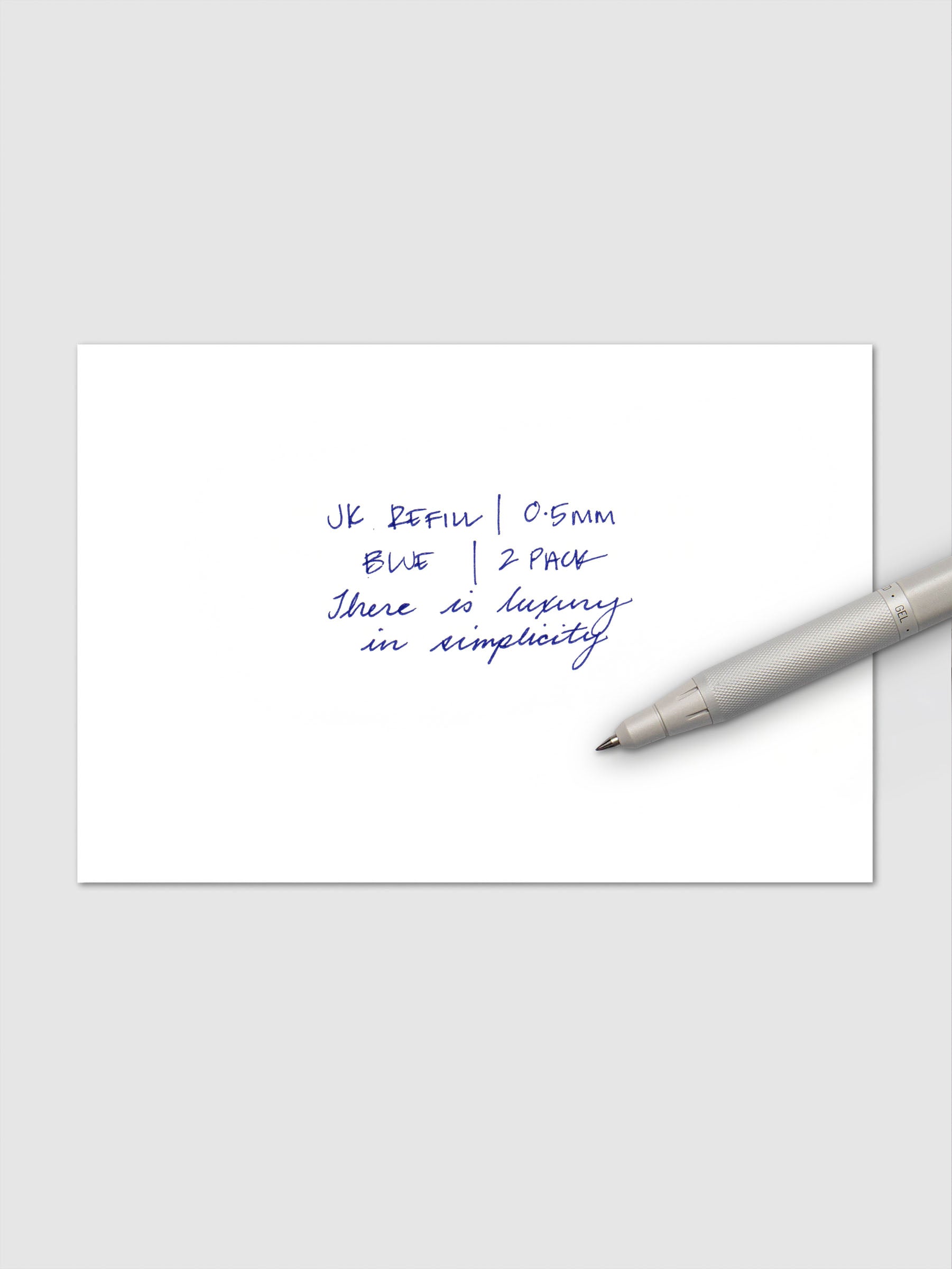 Close-up of handwritten text in blue ink reading "JK Refill | 0.5mm Blue | 2 Pack" followed by "There is luxury in simplicity," with a white Zebra pen resting on the paper.