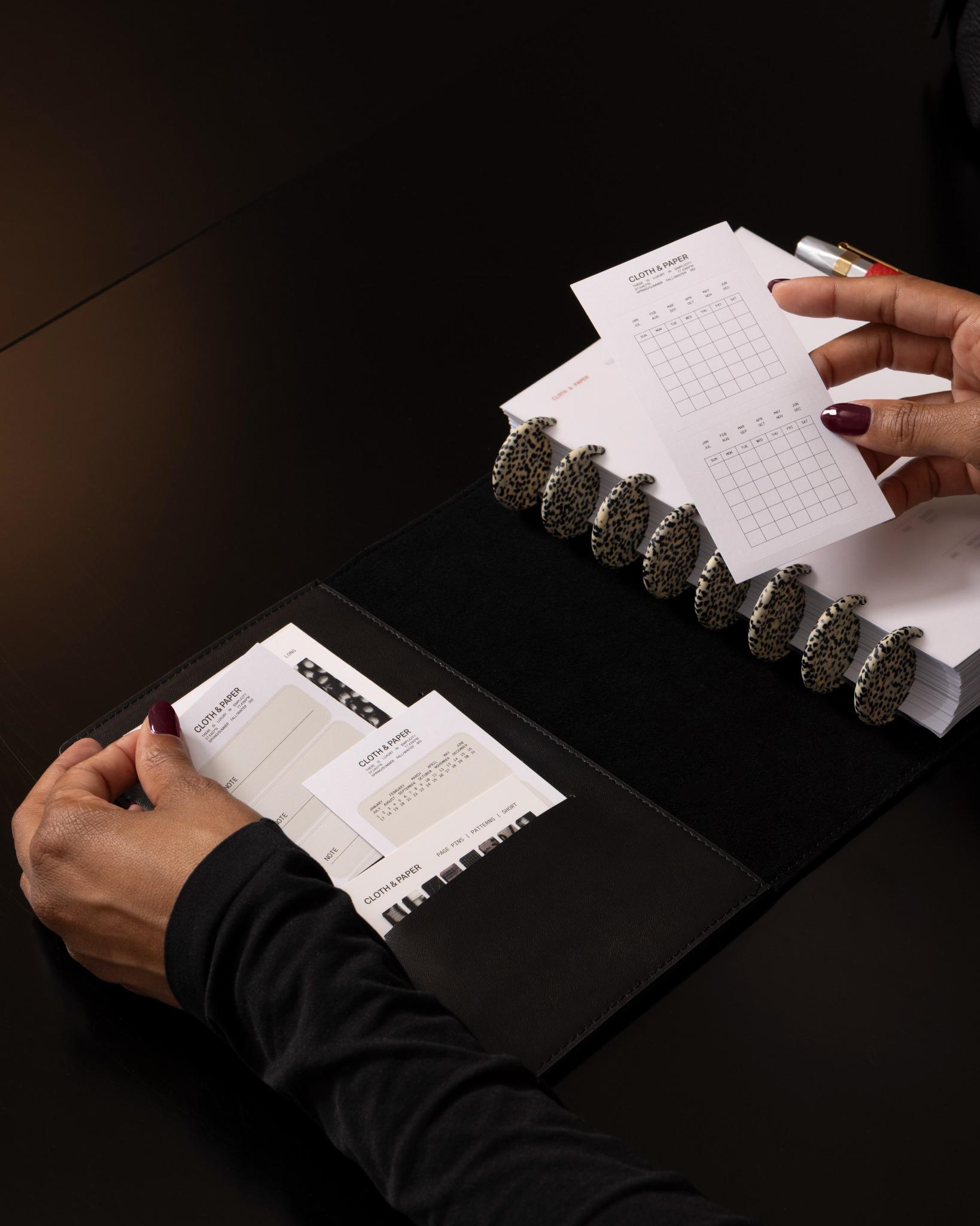 Person placing Cloth & Paper Label Planner Stickers with a calendar design into a black planner folio, with a discbound planner inside the folio