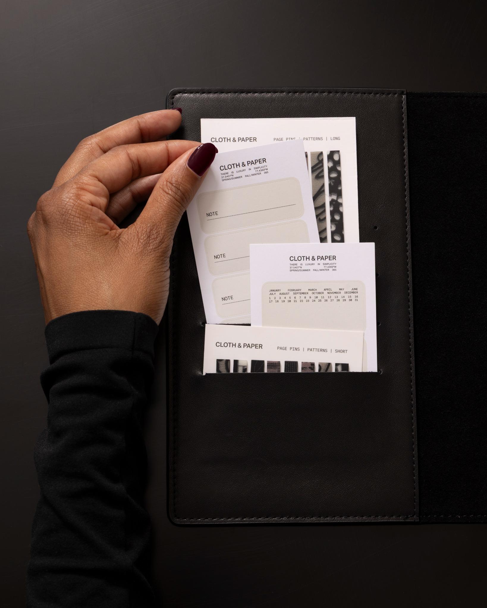 A hand placing label stickers into a black leather agenda pocket, featuring Cloth & Paper stationery including note labels, a mini calendar, and patterned page flags.