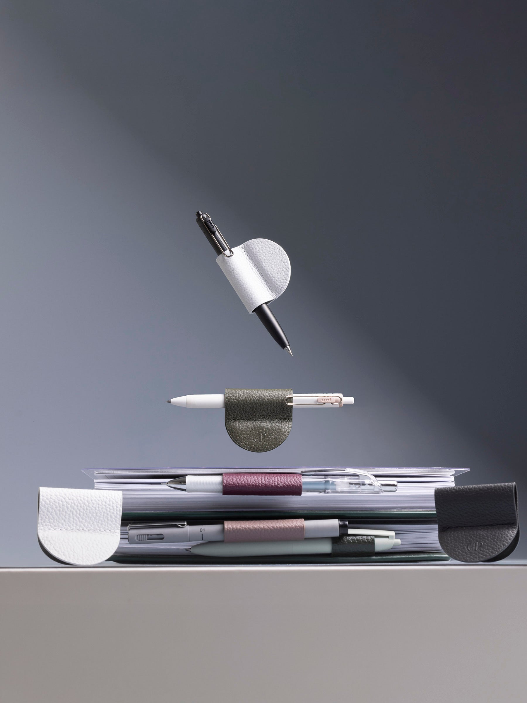 Six clips displayed on a gray background. Two are sitting on a table, facing the viewer. Two are displayed from the side inside of two notebooks. Two are suspended in the air above the notebooks with pens inside of them.