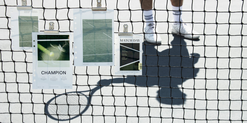 Catch All Clips are holding Luxe Sports Planner Dashboards to a tennis court net.  There is also a person holding a tennis racket behind the net casting a shadow.