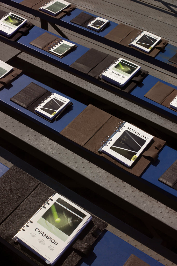 Luxe Sports Planner Dashboards are displayed and layered inside leather planners.  The planners are sitting side by side on steps of bleachers at a tennis court.