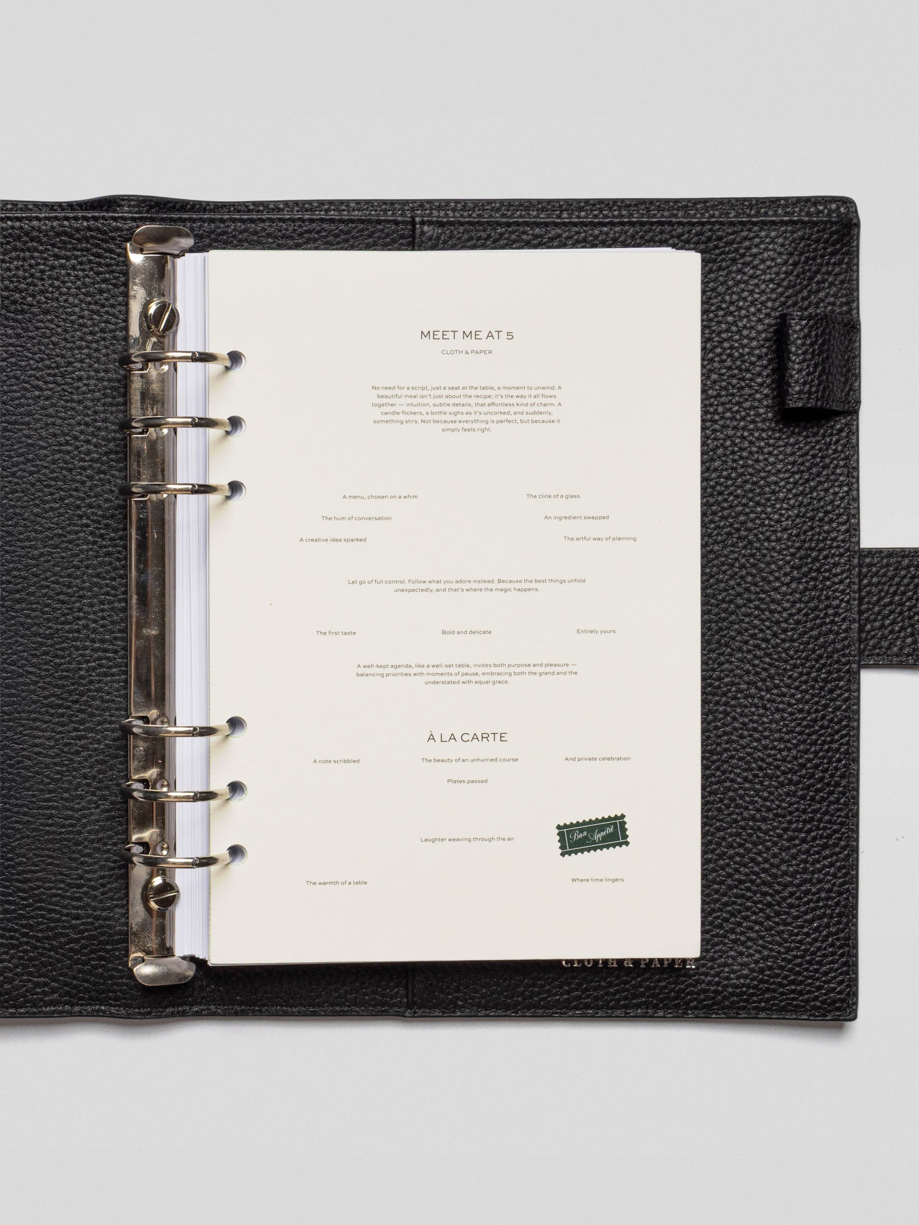 A full-page dashboard titled "Meet Me At 5" is displayed inside a black leather six-ring A5 planner. The cream-colored page features minimalist typography and poetic prompts scattered across the layout.