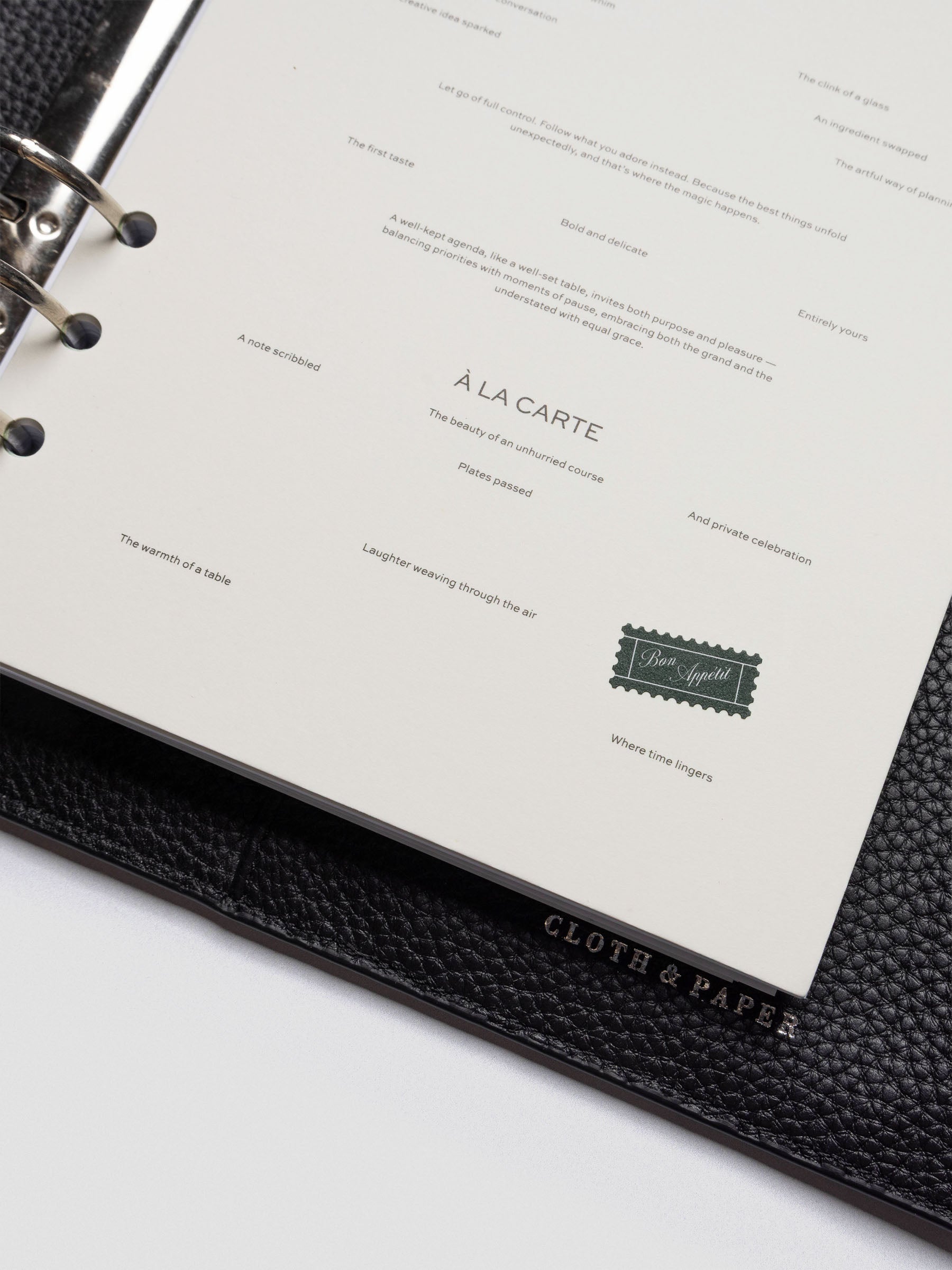 A close-up of the bottom portion of the dashboard, featuring poetic phrases like “The beauty of an unhurried course” and a small green graphic resembling a vintage postage stamp labeled “Bon Appétit.” The edge of the black leather planner embossed with “Cloth & Paper” is also visible.