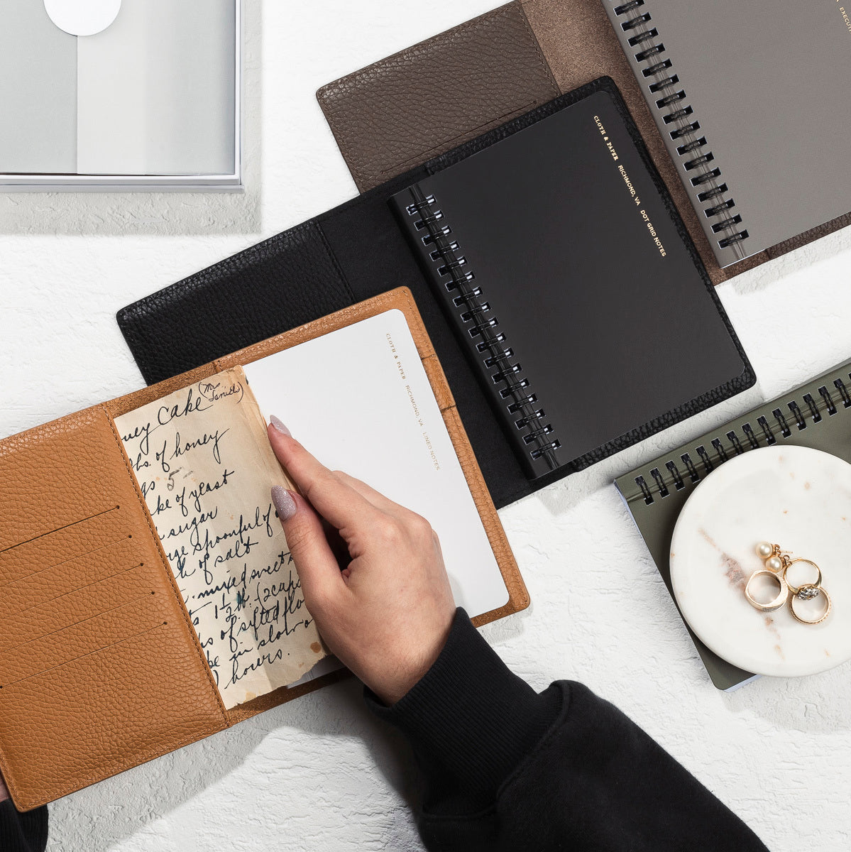 A few Heirloom Leather Folios are opened on a white desk.  A hand is adding some pages of notes to one of the inner pockets.