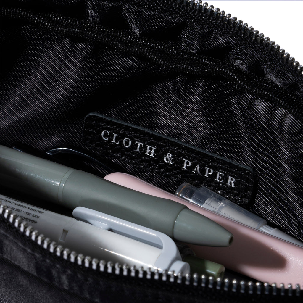 Closeup of Cloth and Paper logo inside of pencil pouch. A selection of pens are stored inside the pouch. Color featured is black.