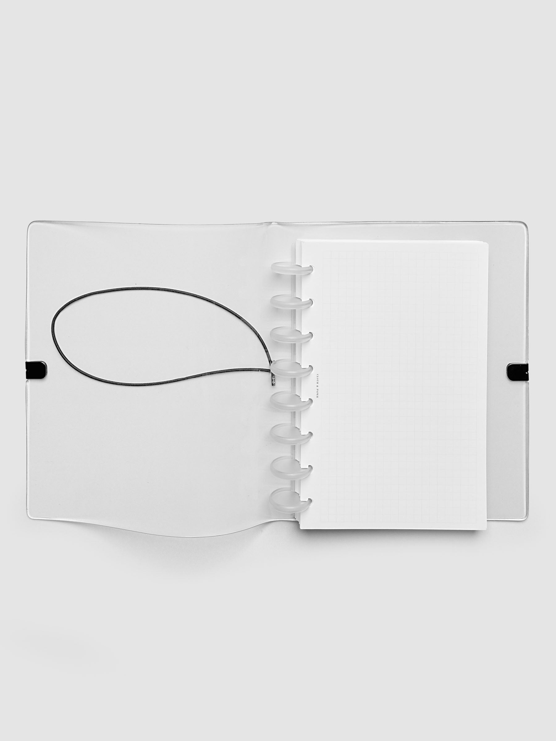 An opened clear plastic notebook cover with a black elastic closure band, with a discbound planner and grid inserts inside, against a light gray background.