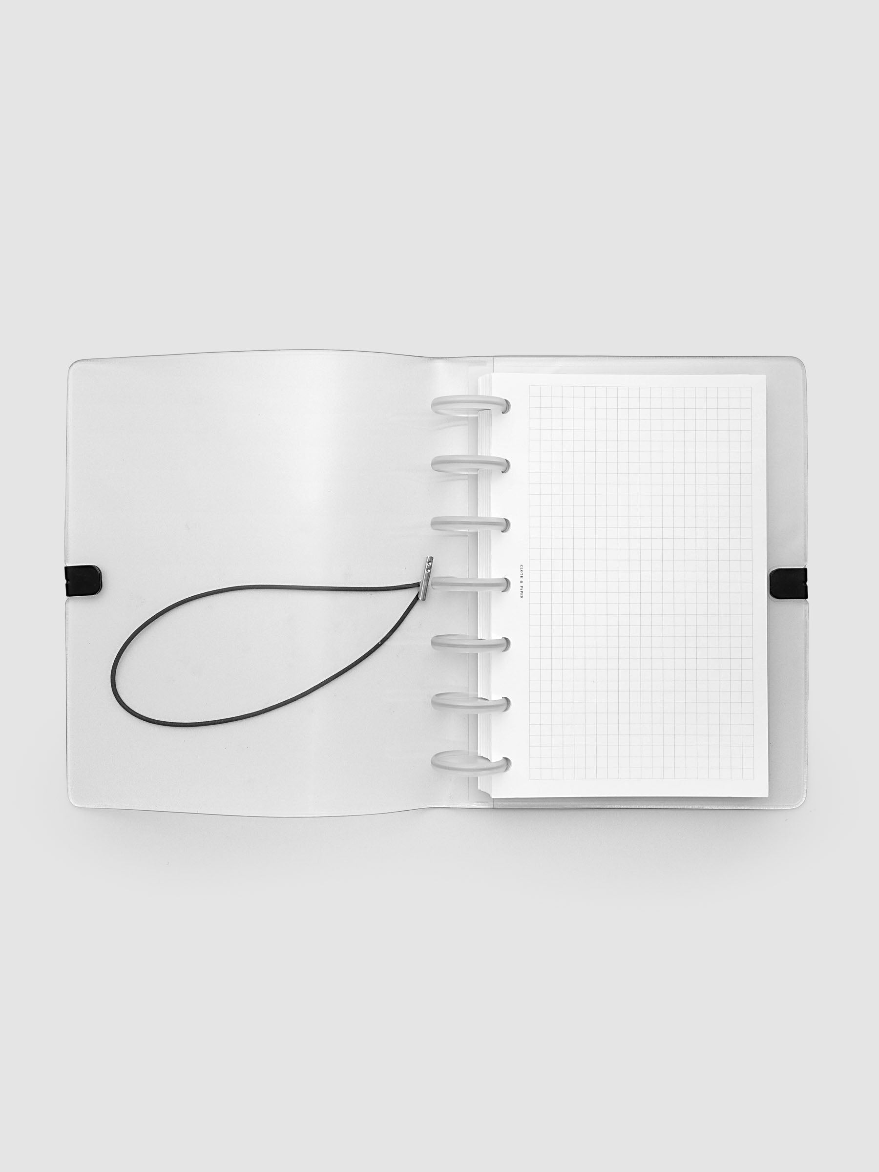 An opened clear plastic notebook cover with a black elastic closure band, with a discbound planner and grid inserts inside, against a light gray background.