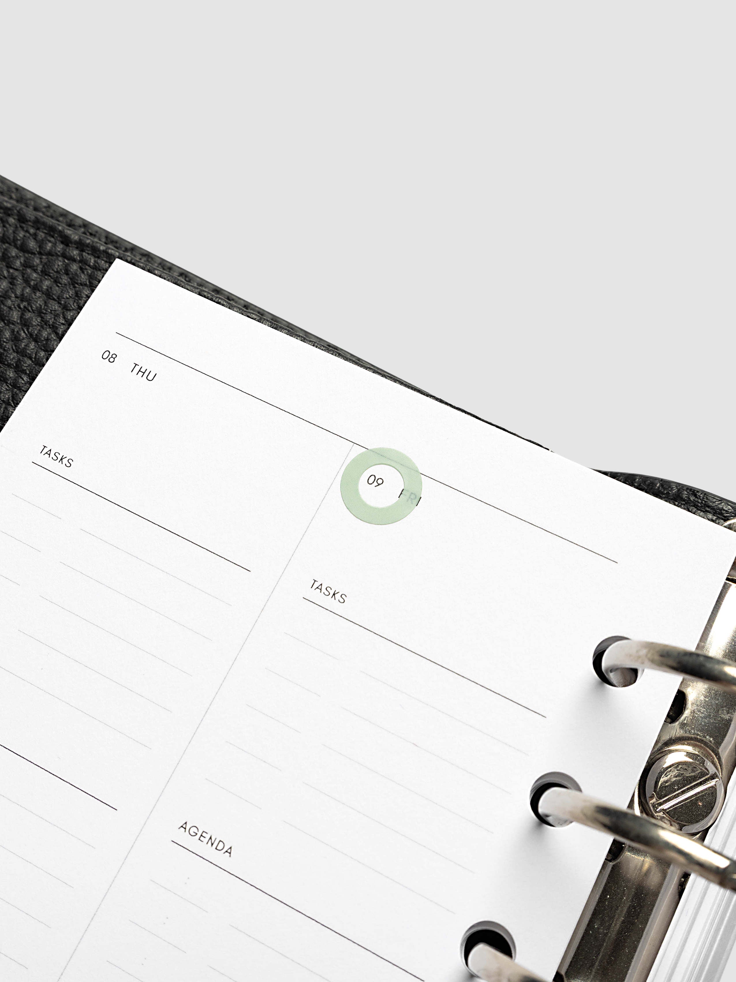 A close-up of a weekly planner page inside a black textured ring binder, showing lined sections for tasks and agenda with a small green sticker highlighting the date.