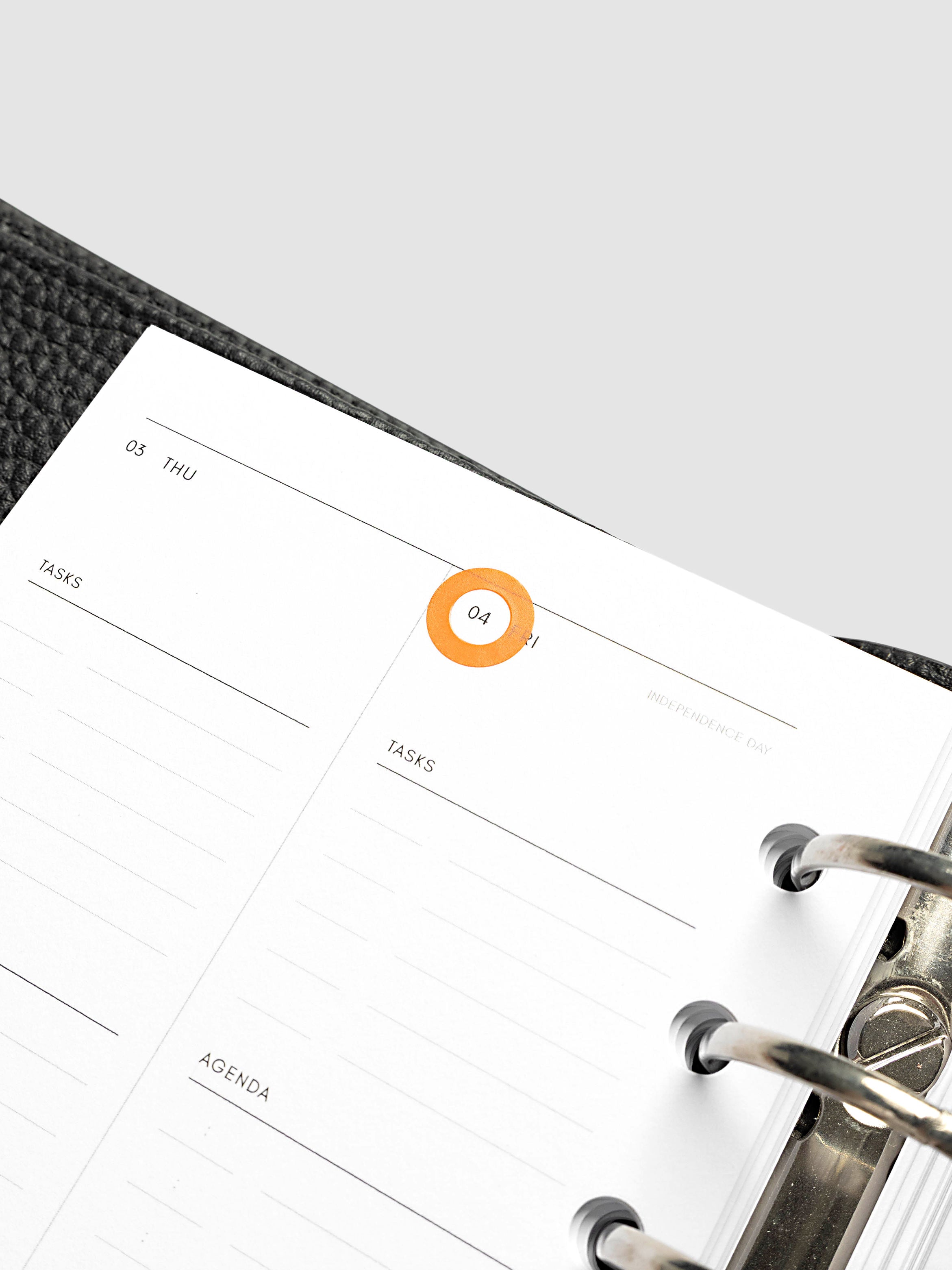 A close-up of a weekly planner page inside a black textured ring binder, showing lined sections for tasks and agenda with a small orange sticker highlighting the date.