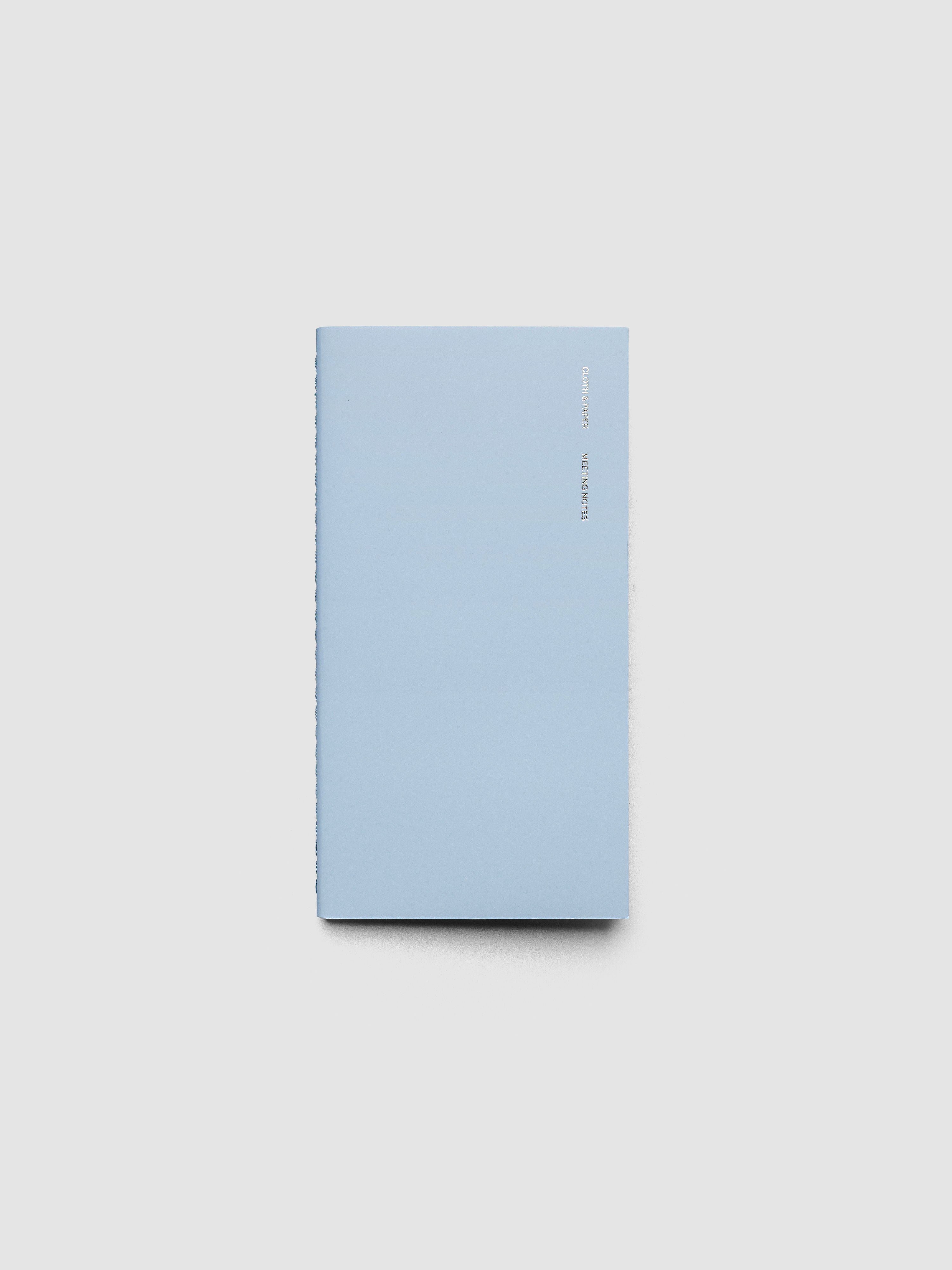 A light blue notebook with a minimalist cover design, featuring a small vertical title in silver foil text on the upper right side, photographed on a neutral light gray background.