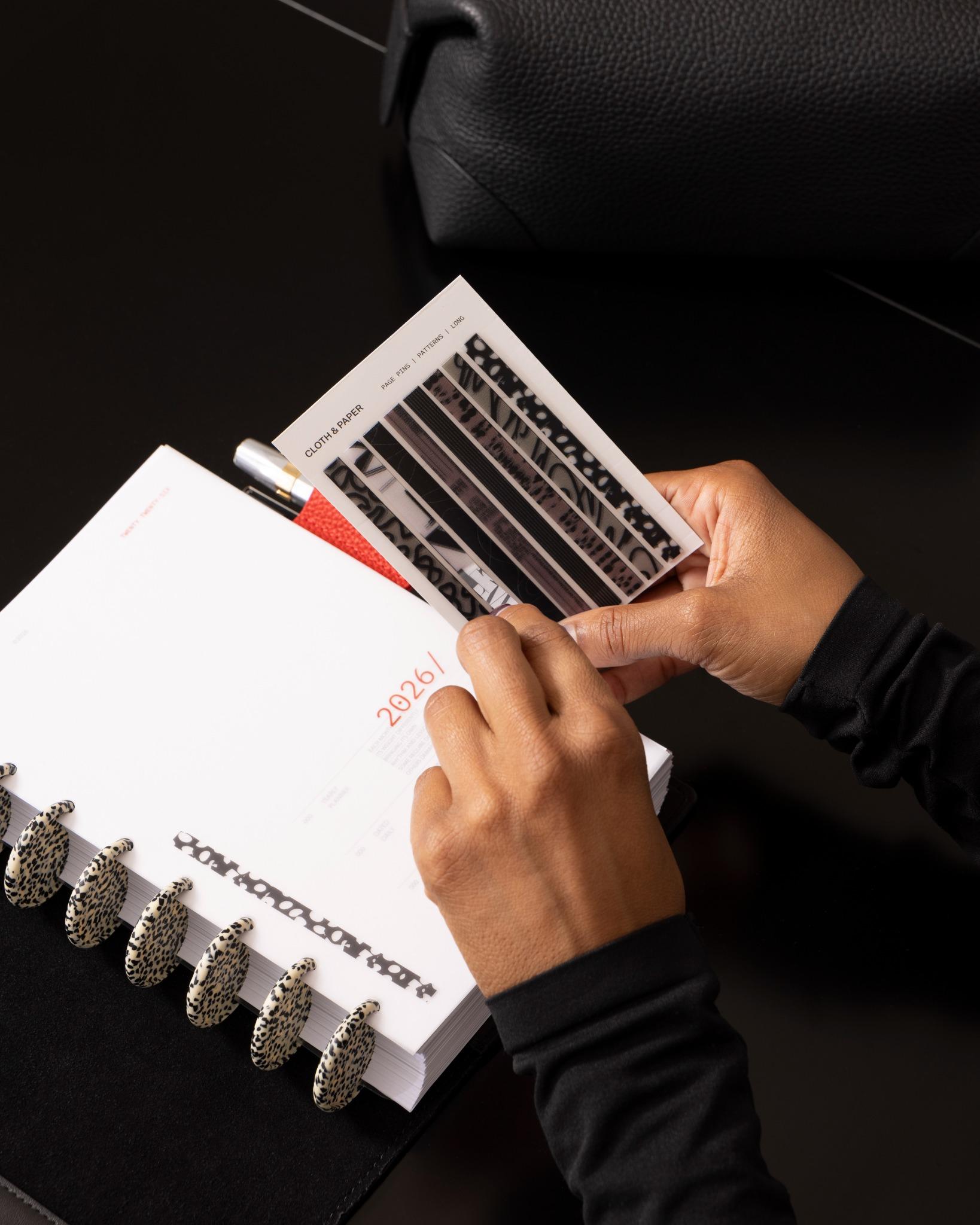 Person placing a sheet of patterned Cloth & Paper Page Pins | Long into a 2026 planner with speckled discs
