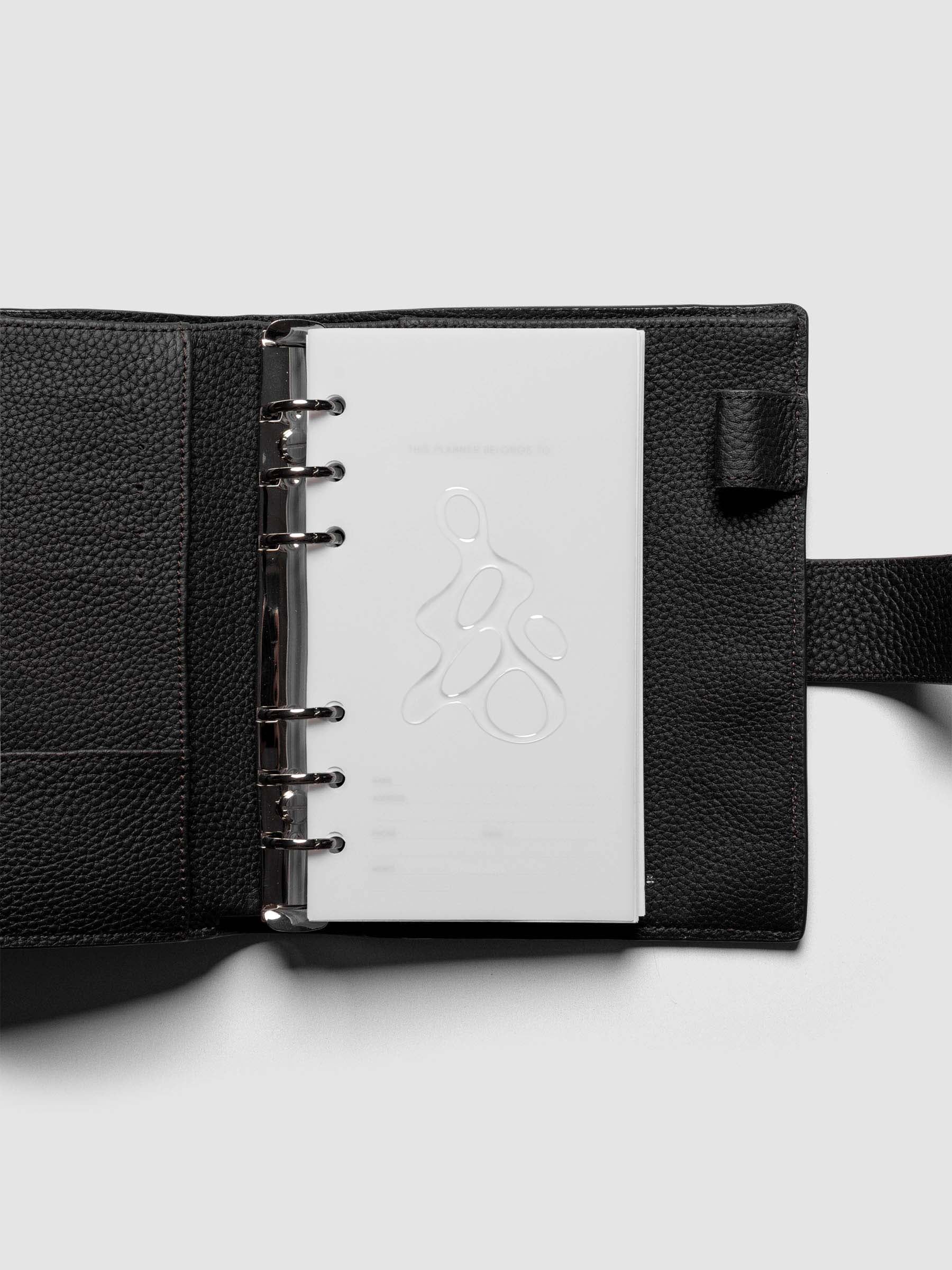 Light grey, semi-transparent, and glossy plastic planner dashboard with organic raised shape design in size Personal shown ringbound in a black leather agenda.
