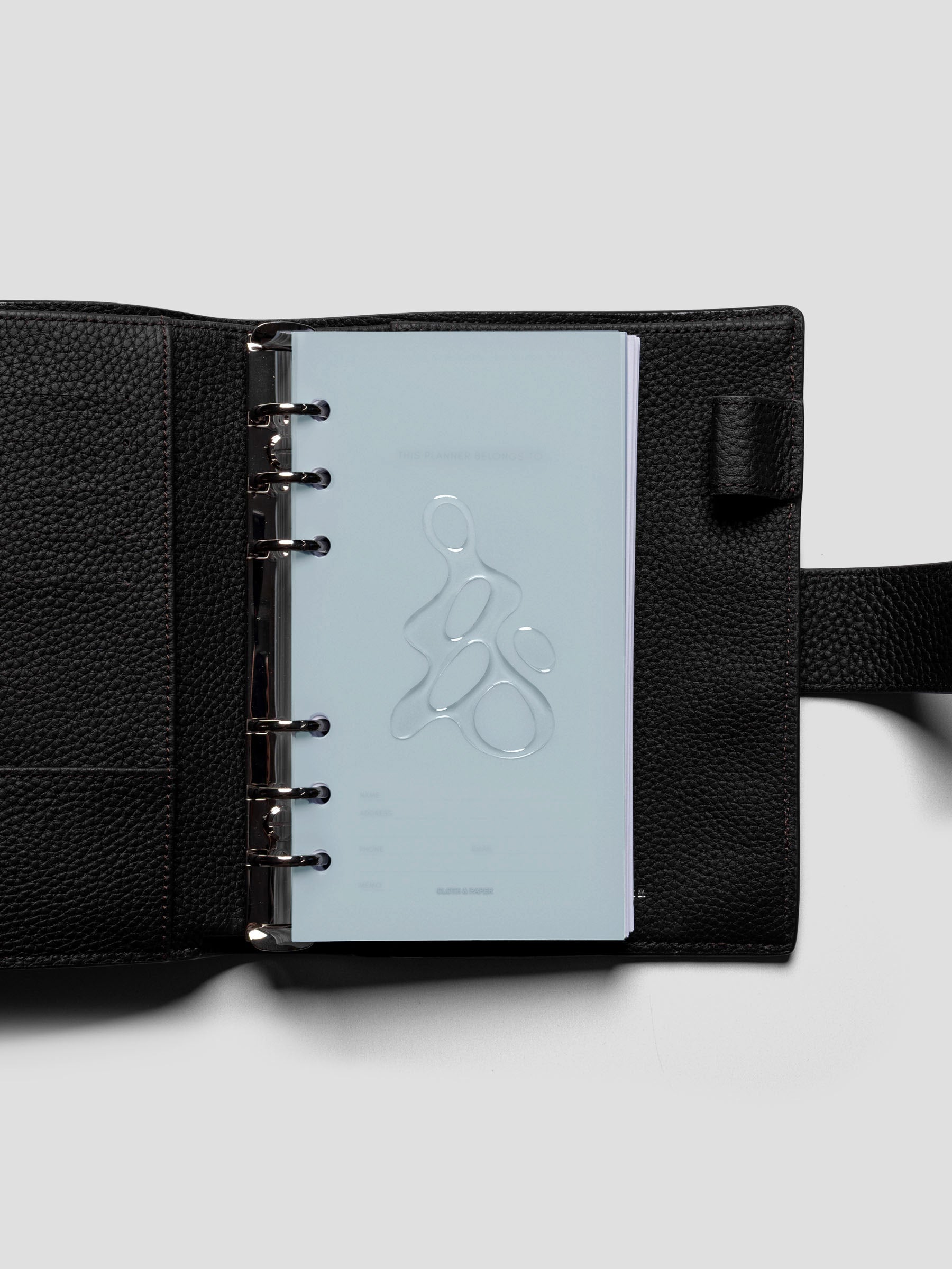 Light blue, semi-transparent, and glossy plastic planner dashboard with organic raised shape design in size Personal on a shown ringbound in a black leather agenda.