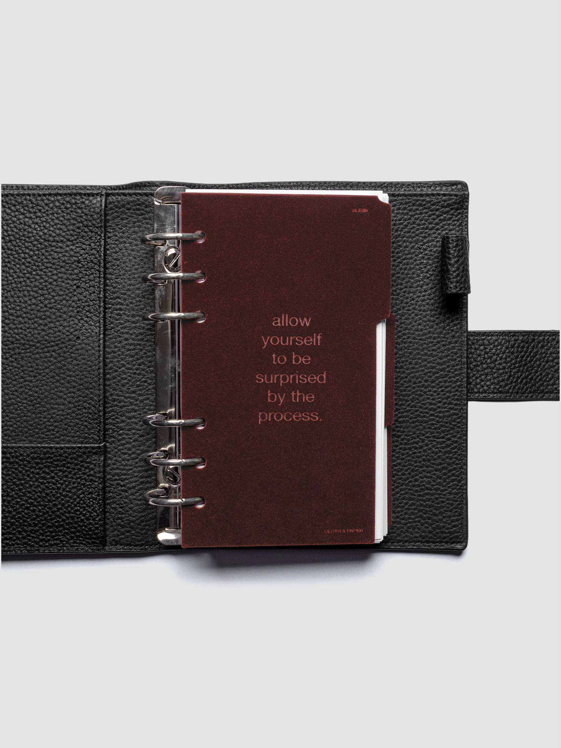 First velvet side tab planners dividers in the color Bordeaux with text deboss design reading "allow yourself to be surprised by the process" in size Personal shown ringbound in a black leather agenda.