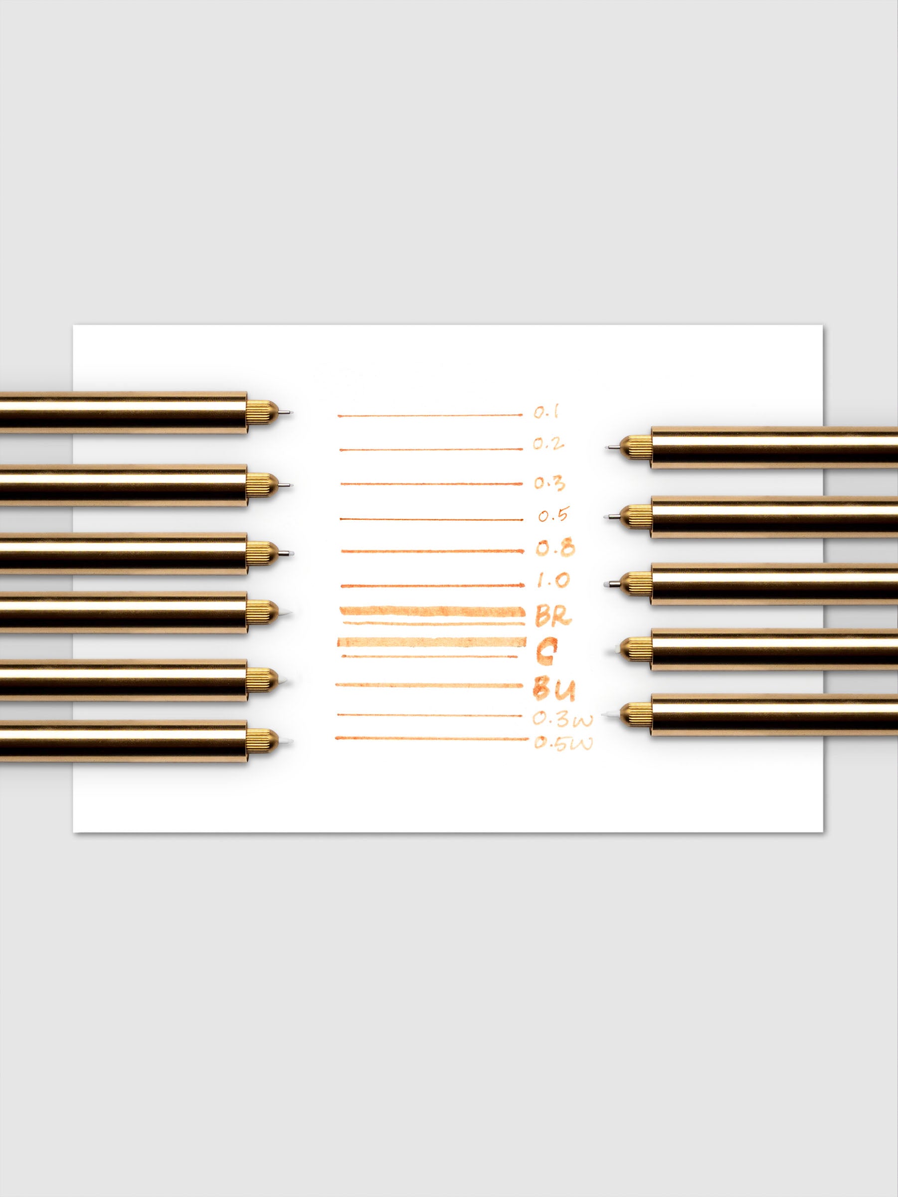 Collection of Lumos Pro Pens in a brass finish showing writing samples on paper with each nib size.
