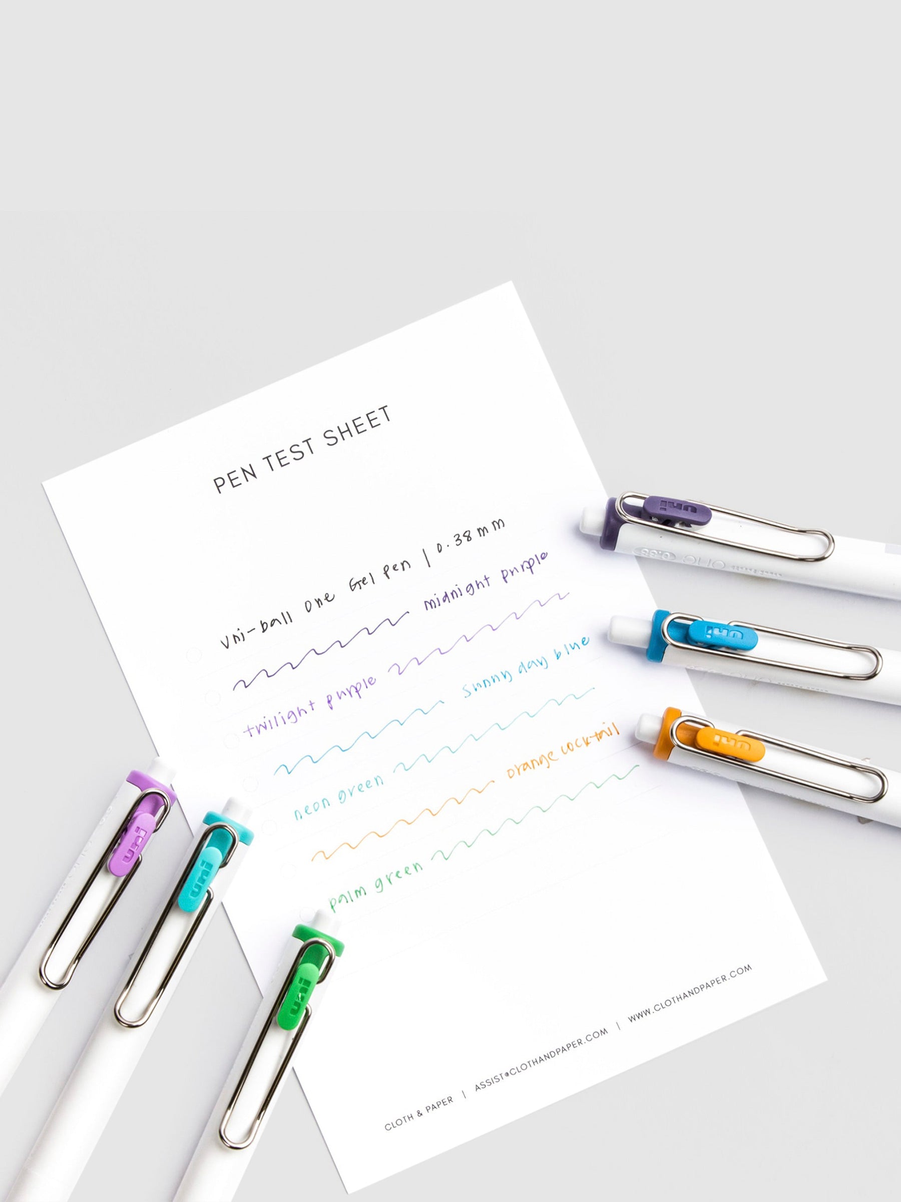 Pen test sheet showing ink samples for Midnight Purple, Neon Green, Orange Cocktail, Palm Green, Sunny Day Blue, and Twilight Purple ink. Six pens are resting on the edges of the pen test sheet.