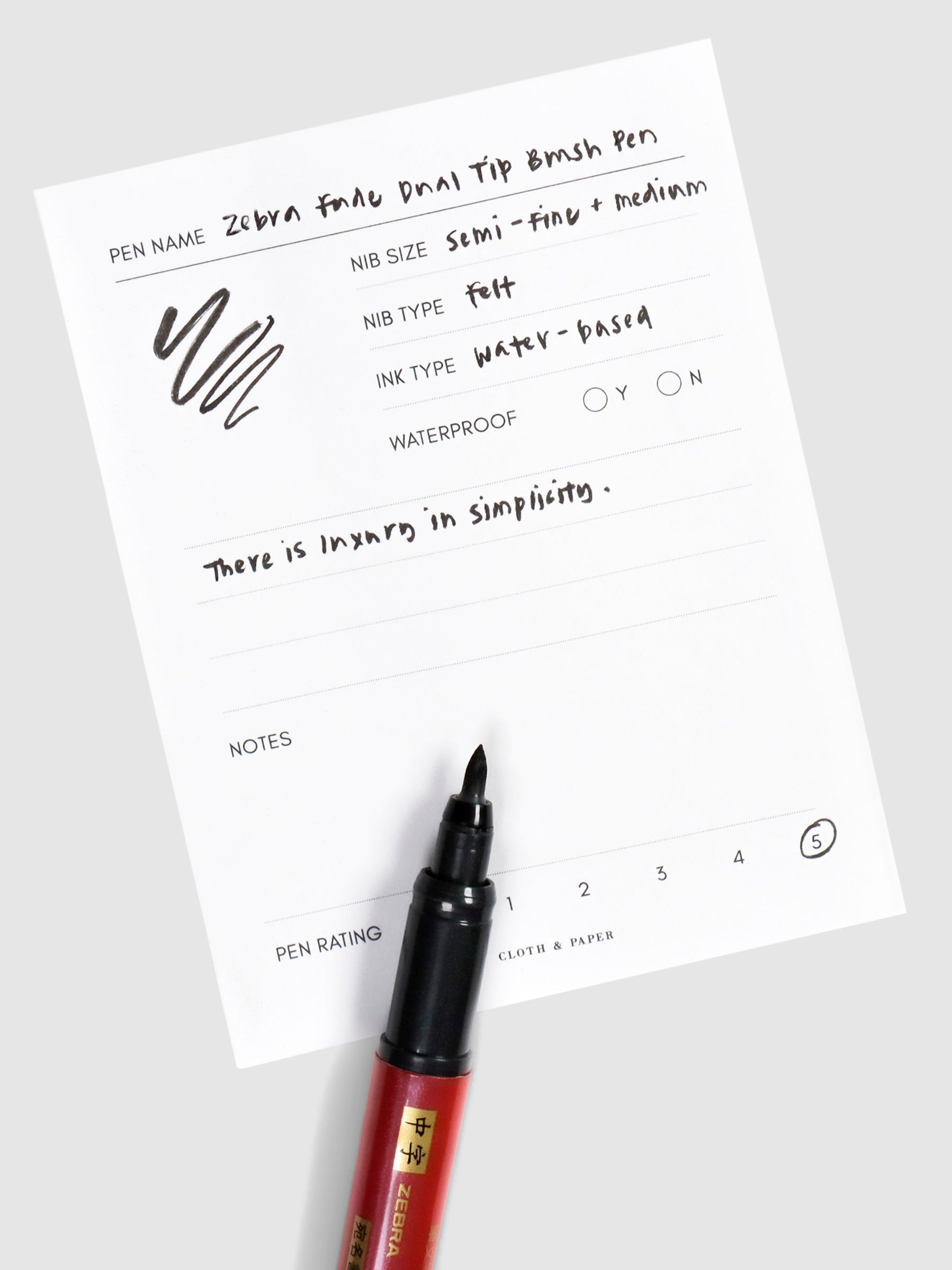 Brush tip nib of pen uncapped, resting on a pen test sheet displaying a writing sample detailing pen specs.