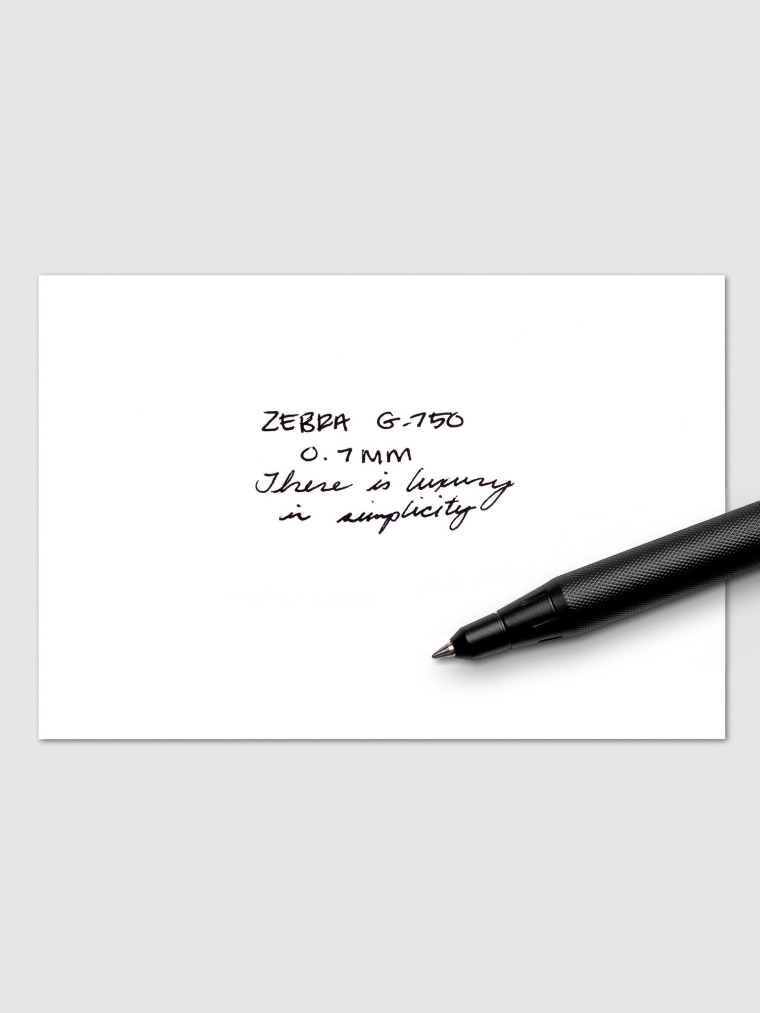 Zebra G-750 Retractable Gel Pen in black barrel shown writing “There is luxury in simplicity” in black ink on white paper, with 0.7 mm tip specification visible.
