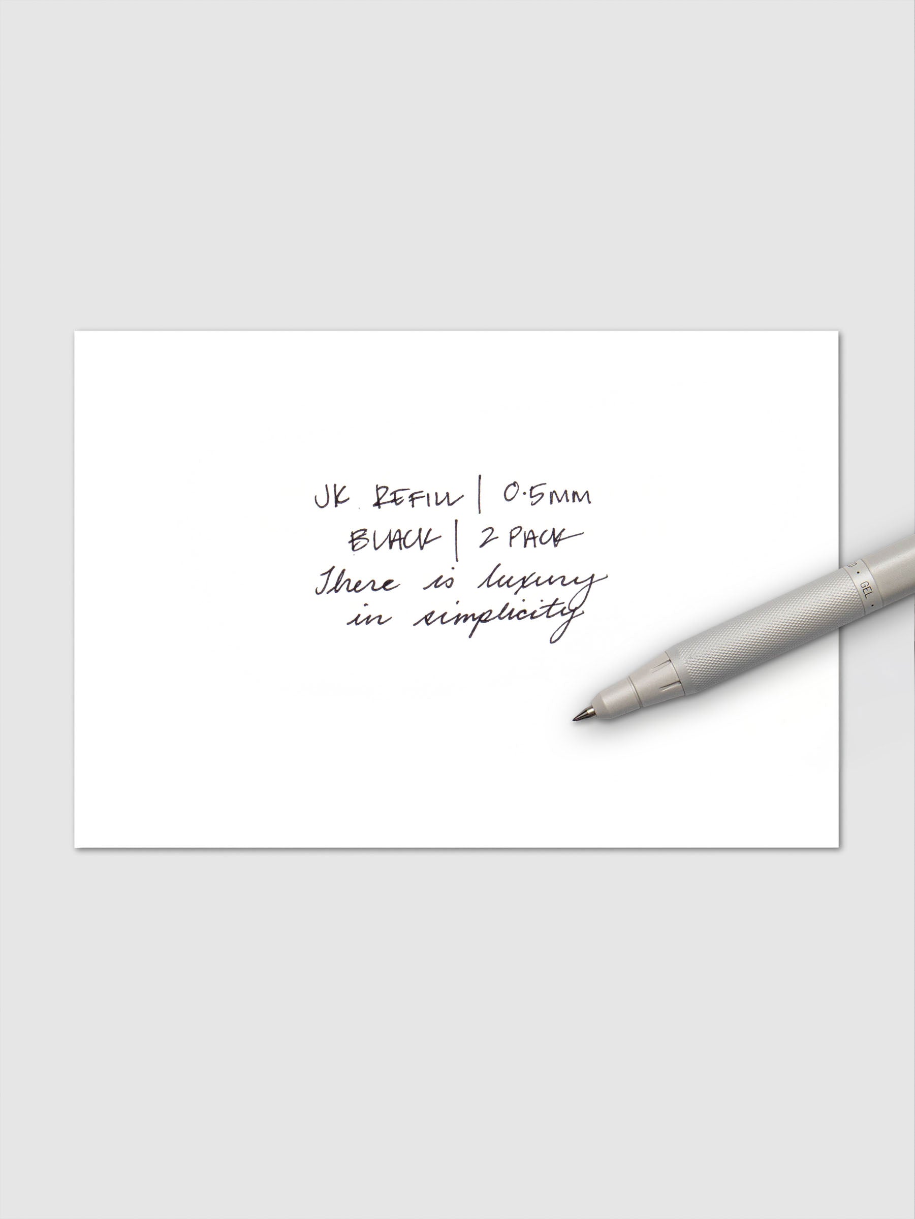 Close-up of handwritten text in black ink reading "JK Refill | 0.5mm Black | 2 Pack" followed by "There is luxury in simplicity," with a white Zebra pen resting on the paper.