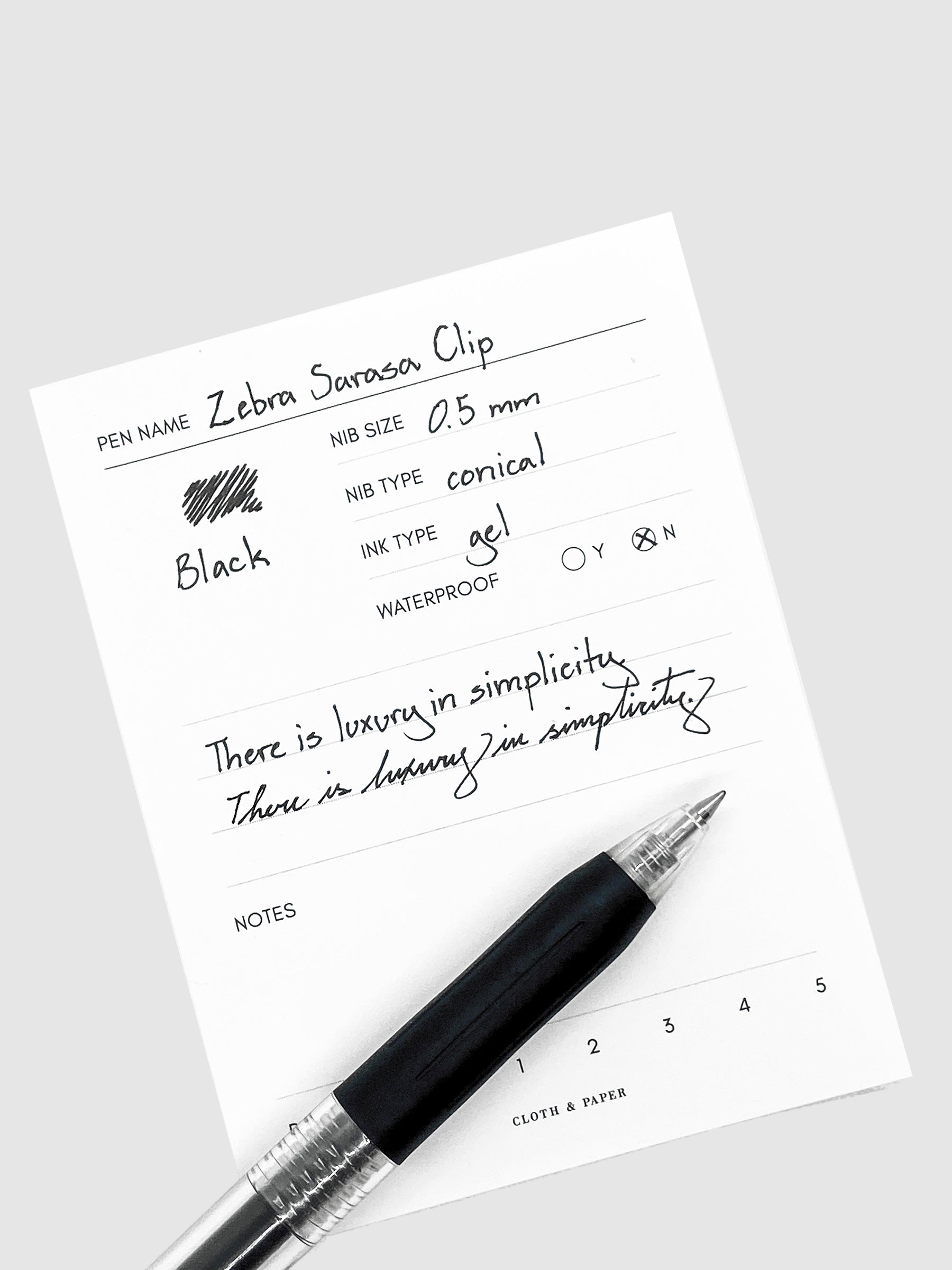 Zebra Sarasa Clip, 0.5 mm, Black, Cloth and Paper. Pen resting on pen test sheet displaying writing sample.