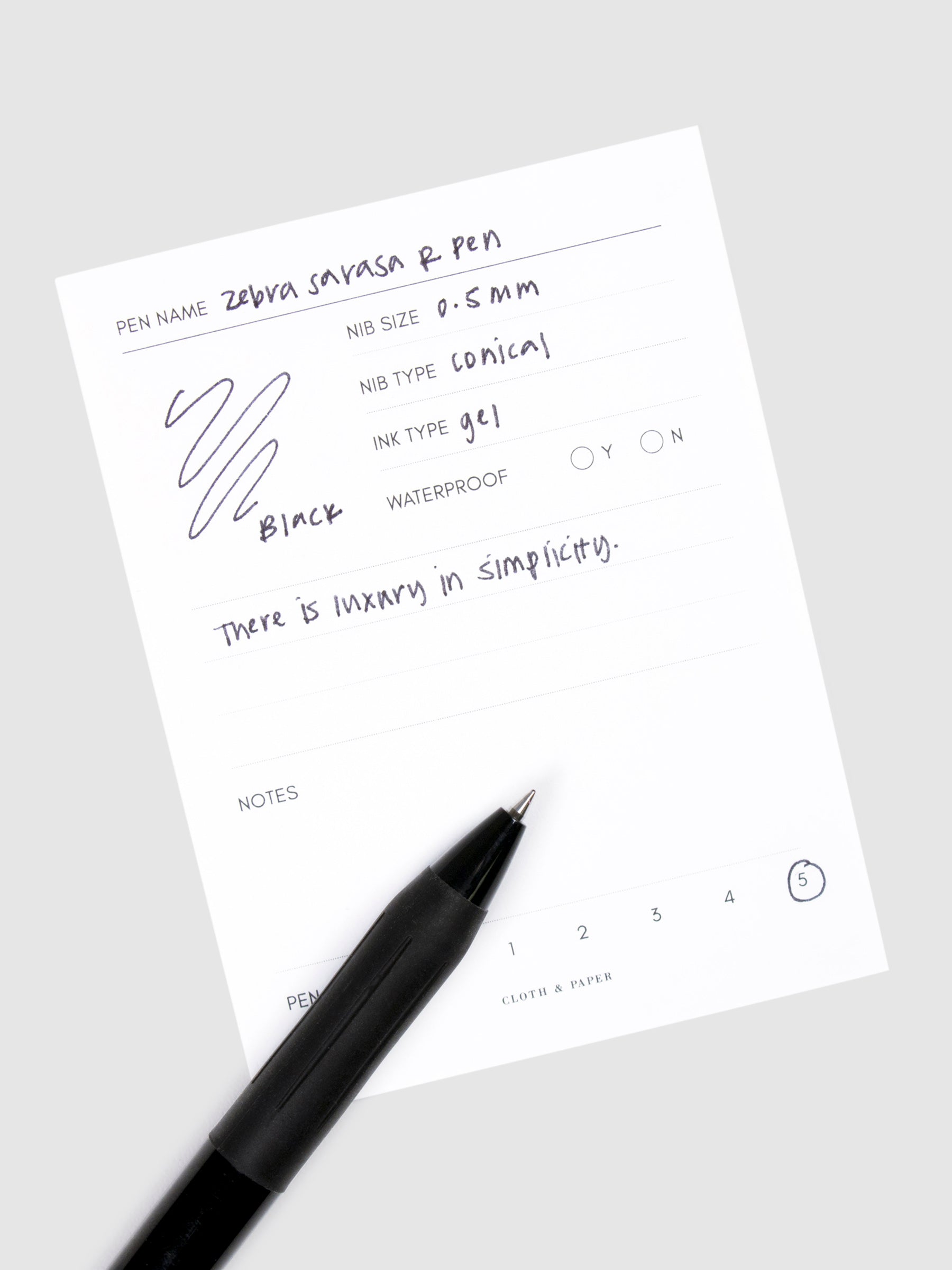 Zebra Sarasa R Pen, 0.5 mm, Black Ink, Black Body, Cloth and Paper. Pen resting on pen test sheet displaying writing sample.