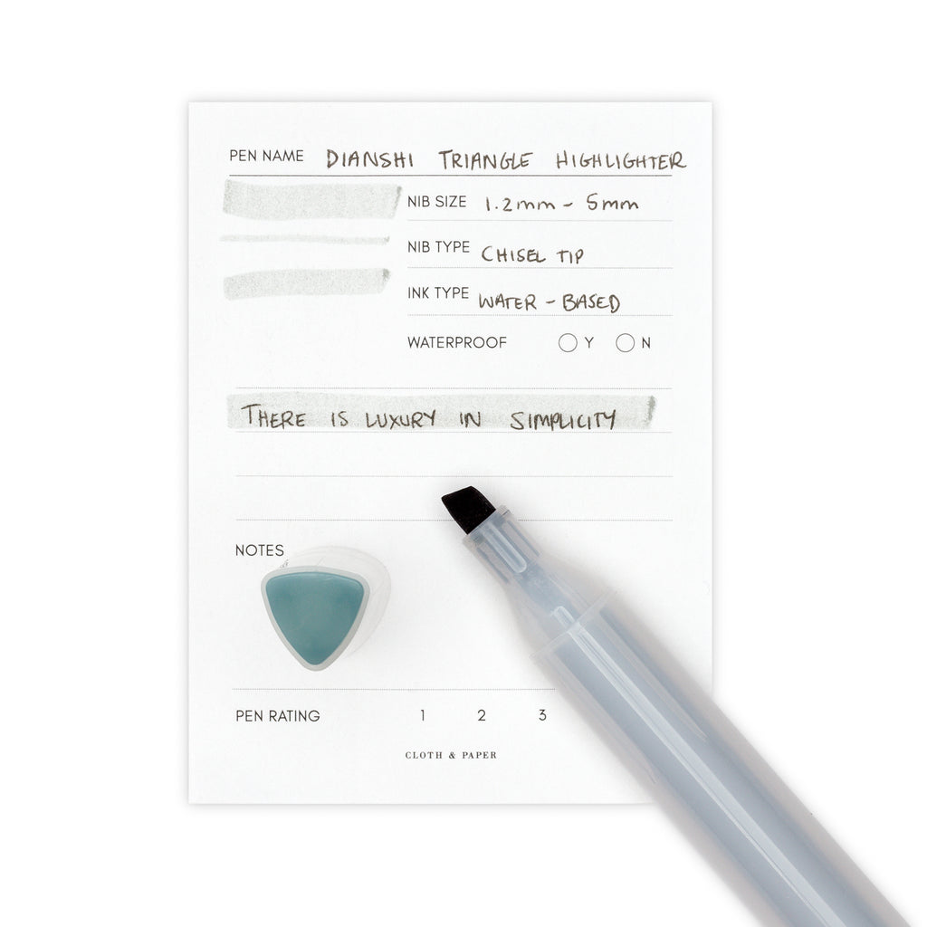 Silver blue highlighter uncapped, resting on a pen test sheet displaying a writing sample detailing the highlighter's specs. The cap stands upright on the bottom left corner of the pen test sheet.