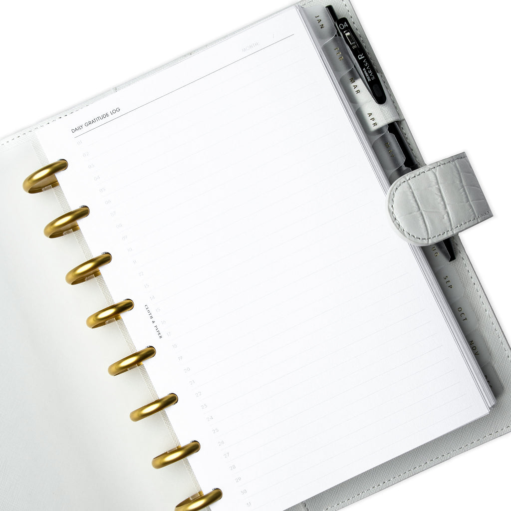 Daily Gratitude Log Planner Inserts styled inside a discbound planner in a white leather agenda cover. The discbound planner has gold rings, and there is black pen tucked into the agenda cover's pen loop.