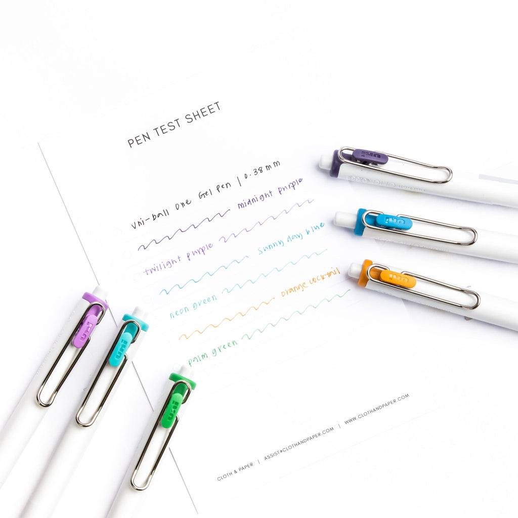 Pen test sheet showing ink samples for Midnight Purple, Neon Green, Orange Cocktail, Palm Green, Sunny Day Blue, and Twilight Purple ink. Six pens are resting on the edges of the pen test sheet.