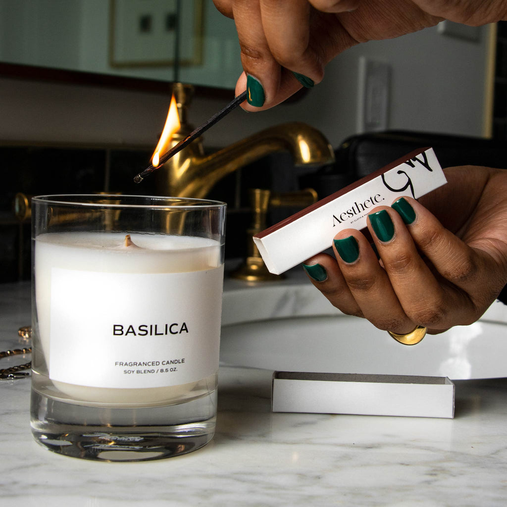 A candle rests on a marble table, about to be lit with a matchstick held by a person with warm brown skin. Their other hand holds a box of Cloth and Paper Aesthete branded matches. 