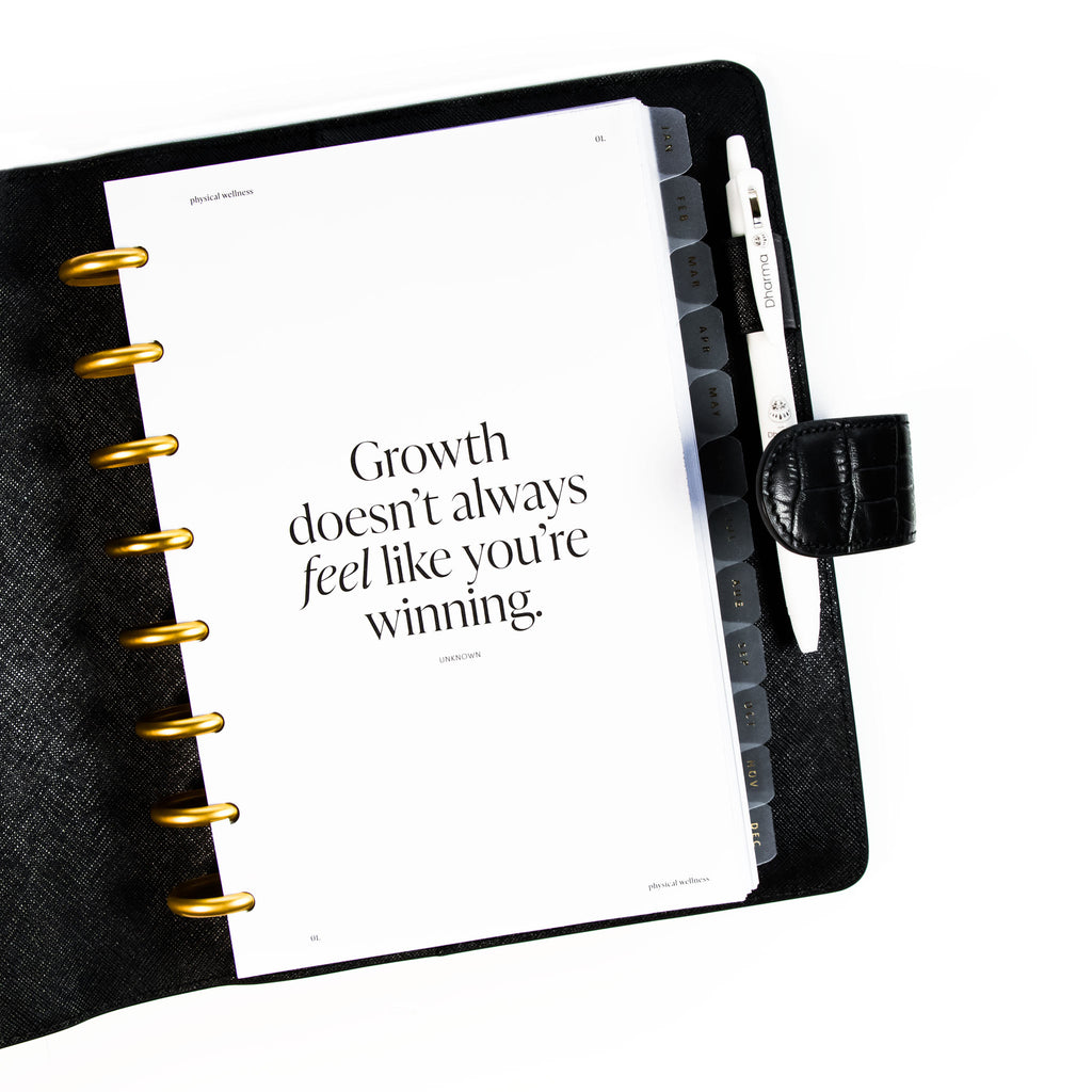 Daily Wellness Planner Insert Bundle styled inside a discbound planner in a black leather cover. The discbound planner has gold discs and the leather cover has a white pen tucked into its pen loop.