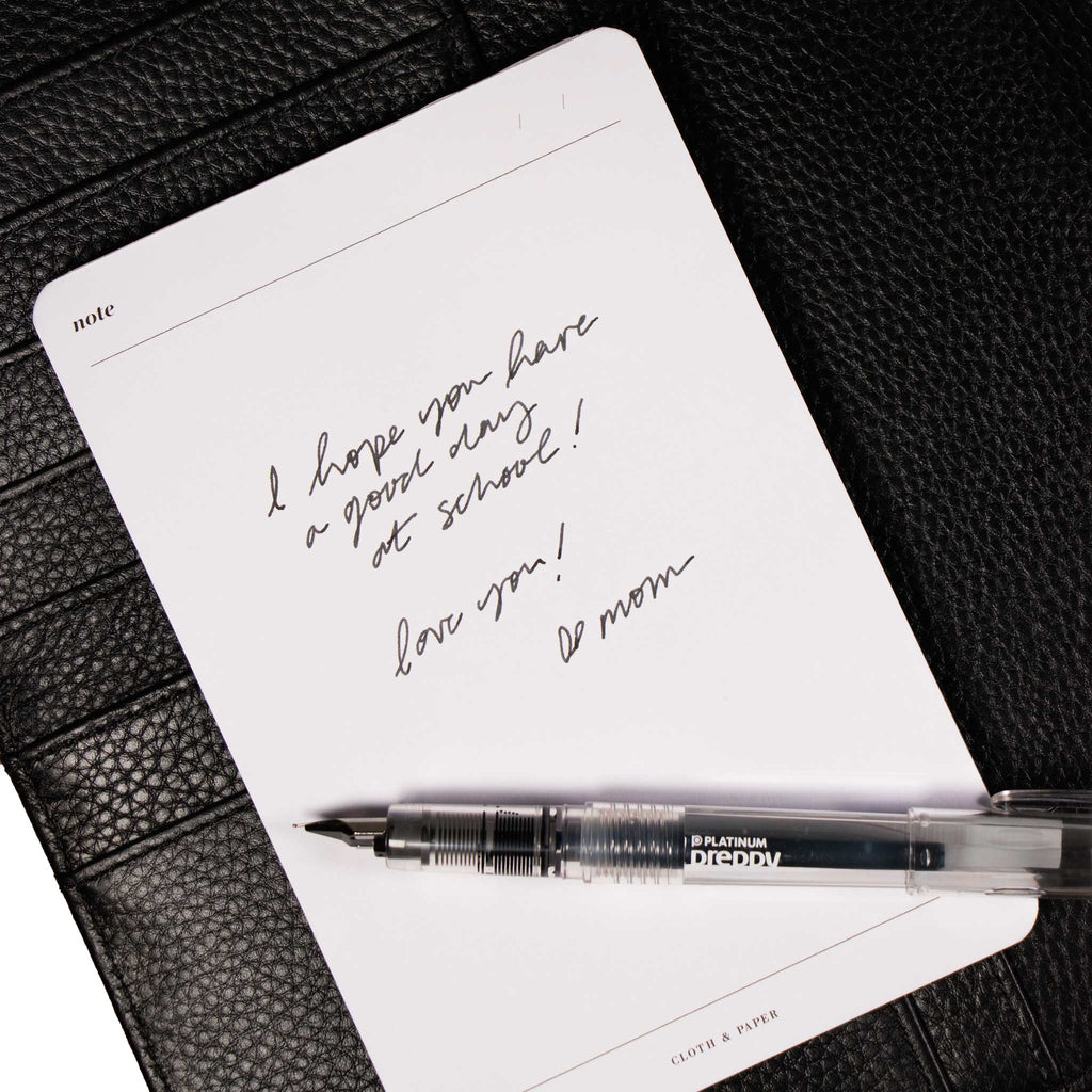 Note Notepad with writing that reads "I hope you have a good day at school! Love you! - Mom" on it. Notepad is styled against a black leather agenda cover with a fountain pen resting on top of it.