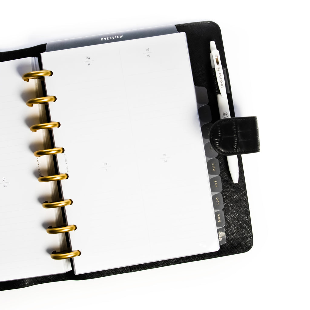 Overview Dry Erase Tab Divider styled inside a discbound planner over undated weekly inserts. The discbound planner has gold discs and rests inside a black leather planner cover.
