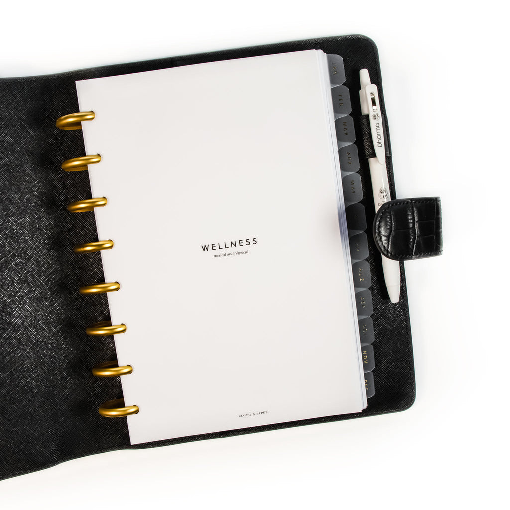 Wellness Section Cover Planner Dashboard styled inside a discbound planner in a black leather planner cover. The discbound planner has gold discs and there is a white pen in the leather cover's pen loop.
