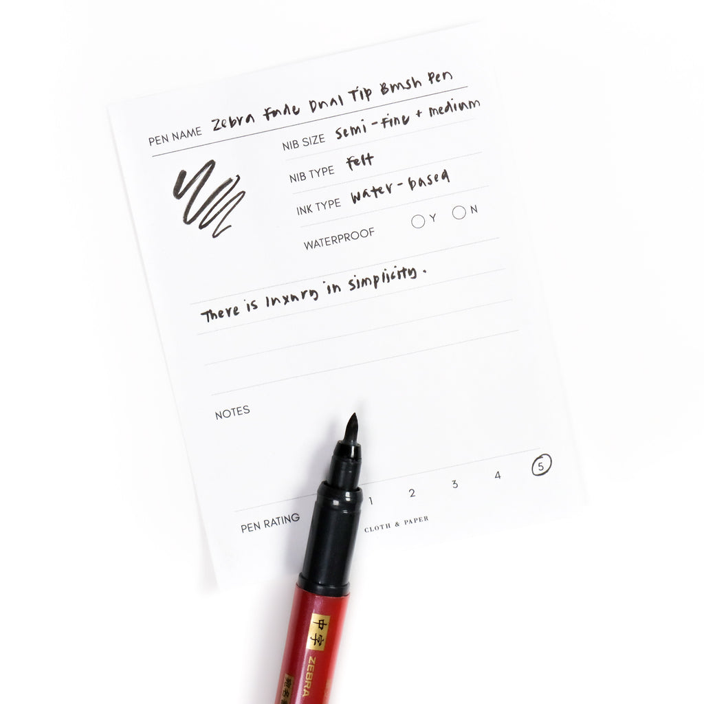 Brush tip nib of pen uncapped, resting on a pen test sheet displaying a writing sample detailing pen specs.